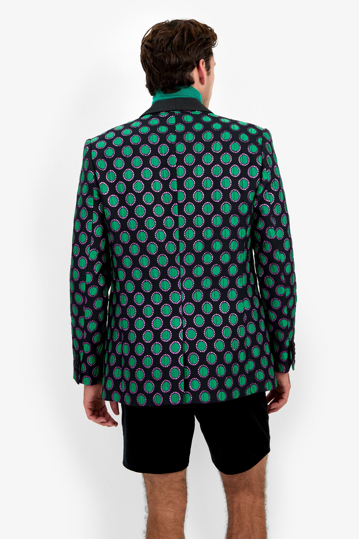 A man stands facing away against a white background, wearing the Alfred Blazer—a dark patterned blazer with green and purple ovals—paired with a green-collared shirt and black shorts.