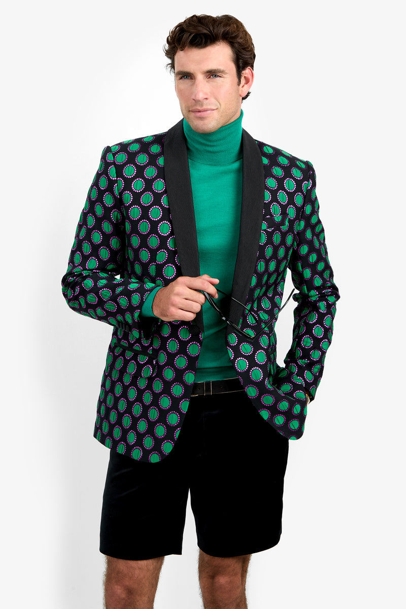 A man in a green turtleneck, the Alfred Blazer with black lapels and patterned design, and black shorts holds glasses and stands against a white background.