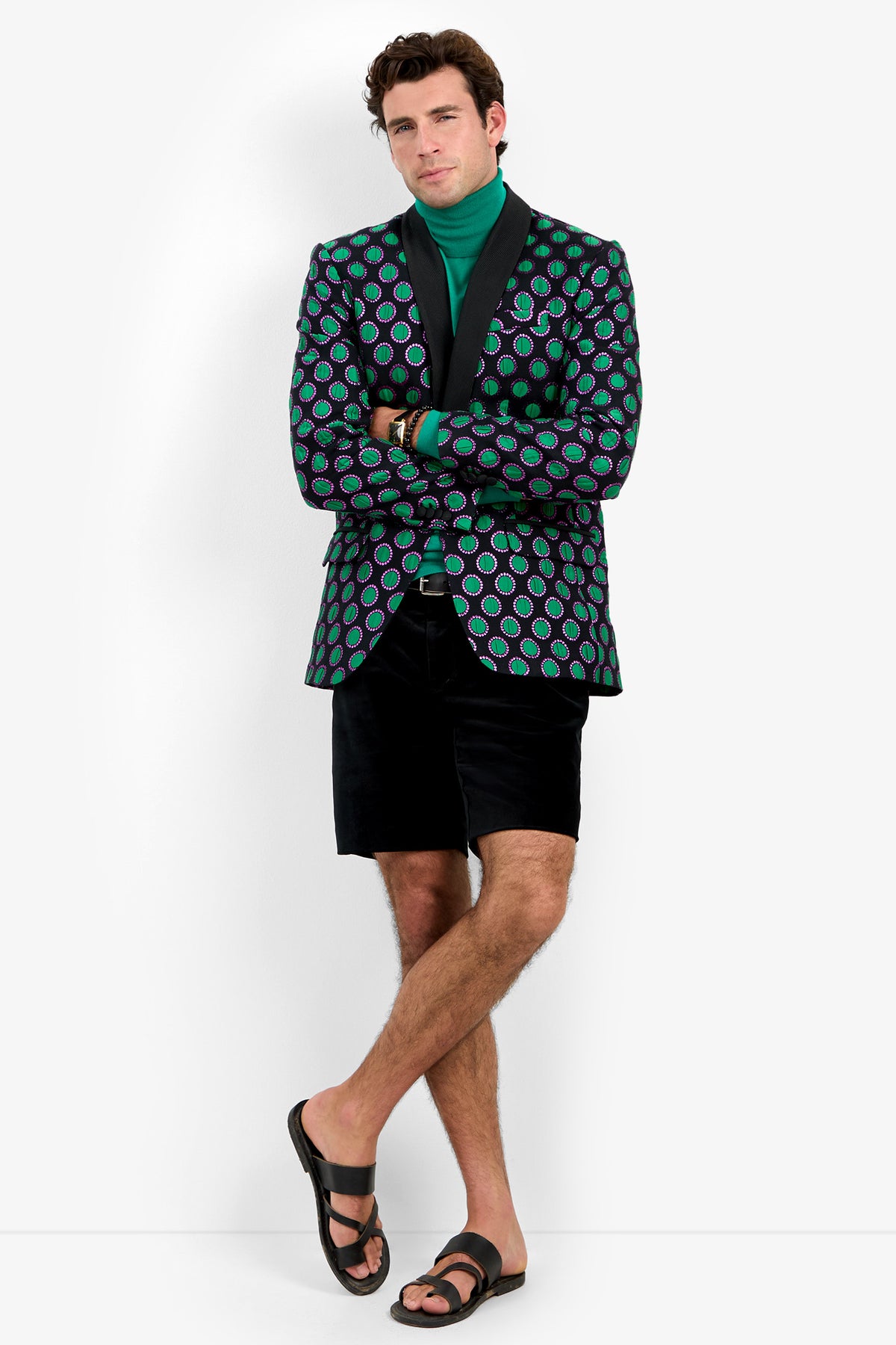 A man wearing the Alfred Blazer, styled with a green turtleneck, black shorts, and sandals, stands with his legs crossed and looks relaxed against a plain white background.