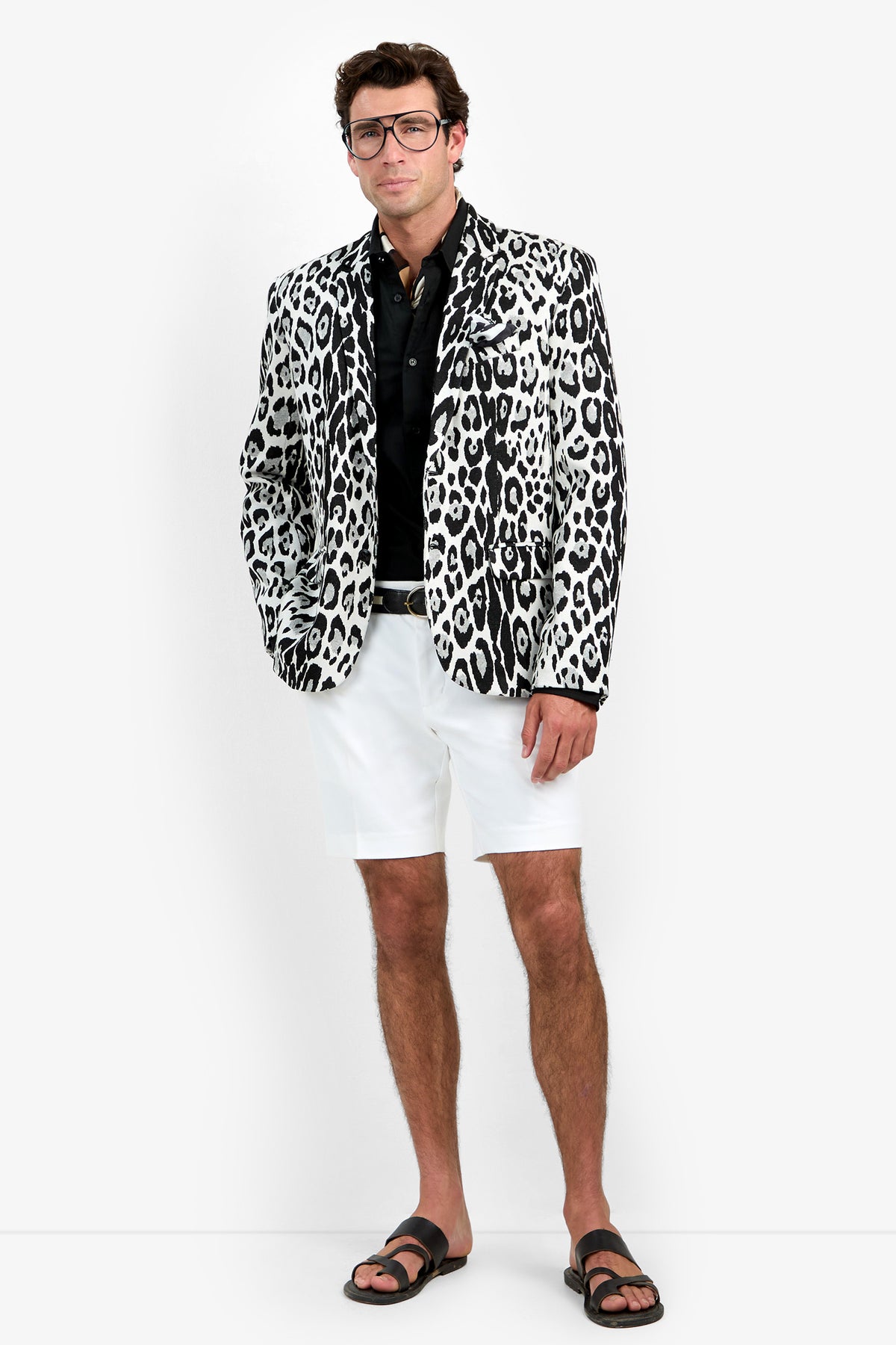 A man wearing glasses, a black shirt, white shorts, black sandals, and the Thurston Blazer stands against a plain white background.