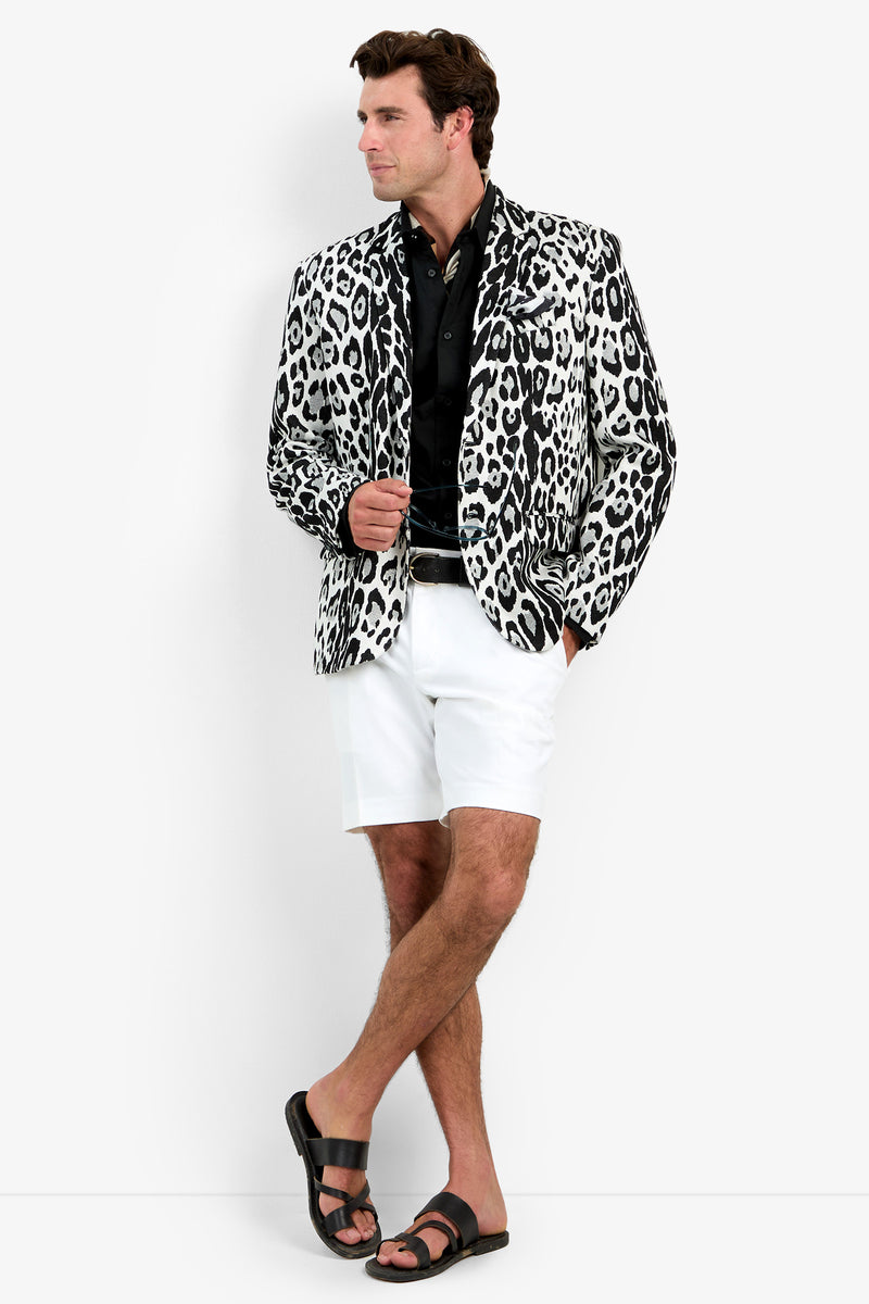A man wearing the Thurston Blazer stands against a white background with one leg crossed, looking to the side. He pairs the blazer with a black shirt, white shorts, and black sandals.