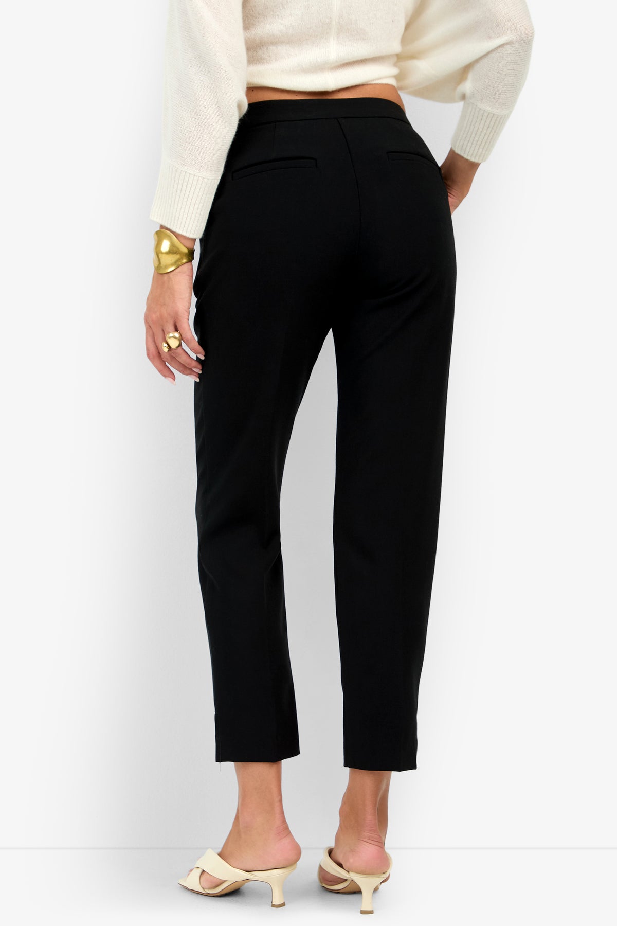 Back view of the black ARNO PANT high-waisted ankle trousers, paired with a cream sweater and cream-colored kitten heel mules.