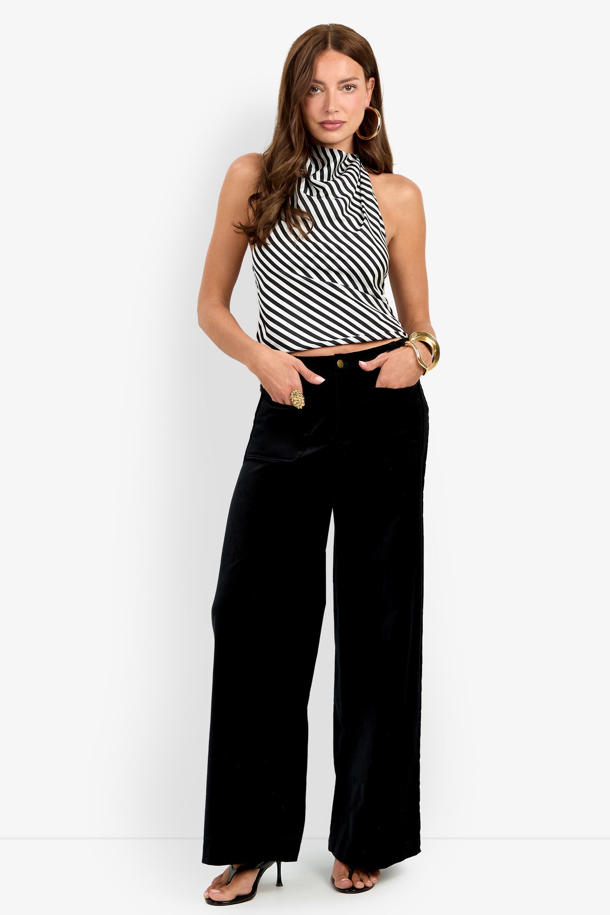 Wearing the Talamone Pant, a woman styles it with a black and white striped sleeveless turtleneck top, black heeled sandals, gold hoop earrings, and chunky gold bracelets as she stands against a plain background.