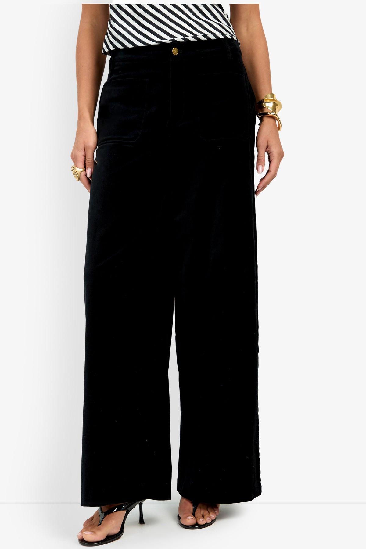 A person models the Talamone Pant—a high-waisted, wide-leg black style—paired with a striped top, gold bangles, rings, and black heeled sandals against a white background. Only the lower half of their body is shown.