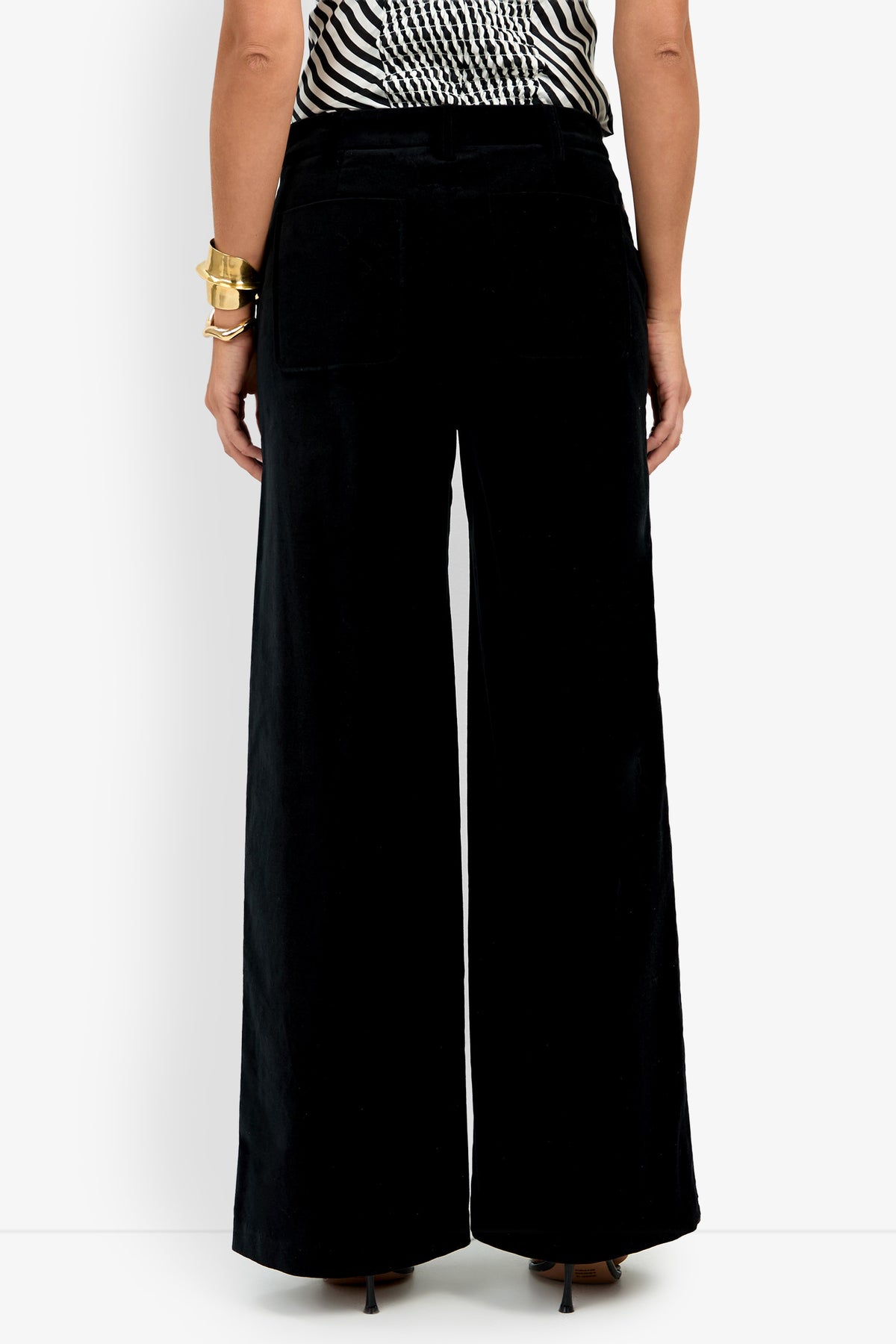 Wearing the Talamone Pant in a wide-leg black style, this individual pairs them with a black-and-white striped sleeveless top and gold bracelets on their left wrist for a stylish, modern look.