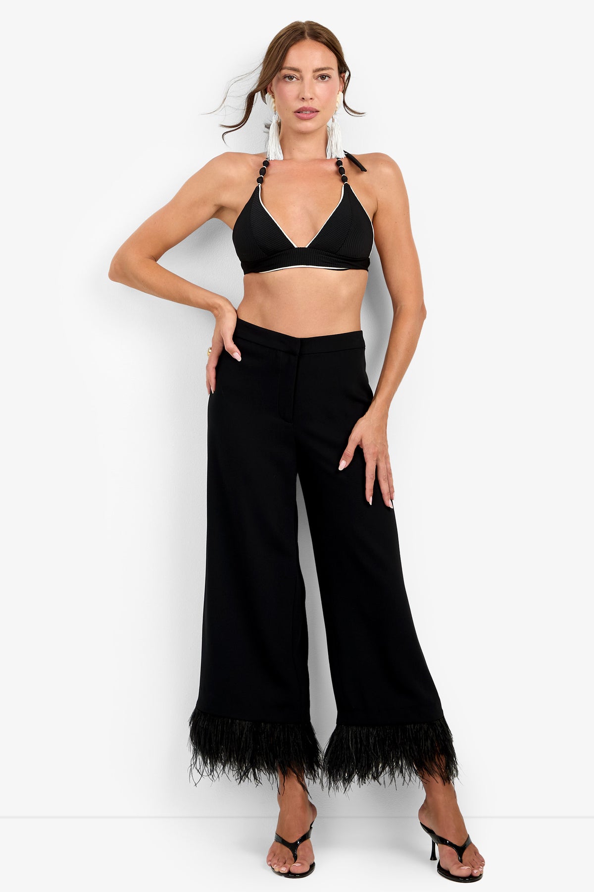 A woman poses against a plain white background wearing the Amarie Pant in black—a wide-leg silhouette featuring feather trim at the hem—styled with a black halter bikini top, black heeled sandals, and statement earrings.