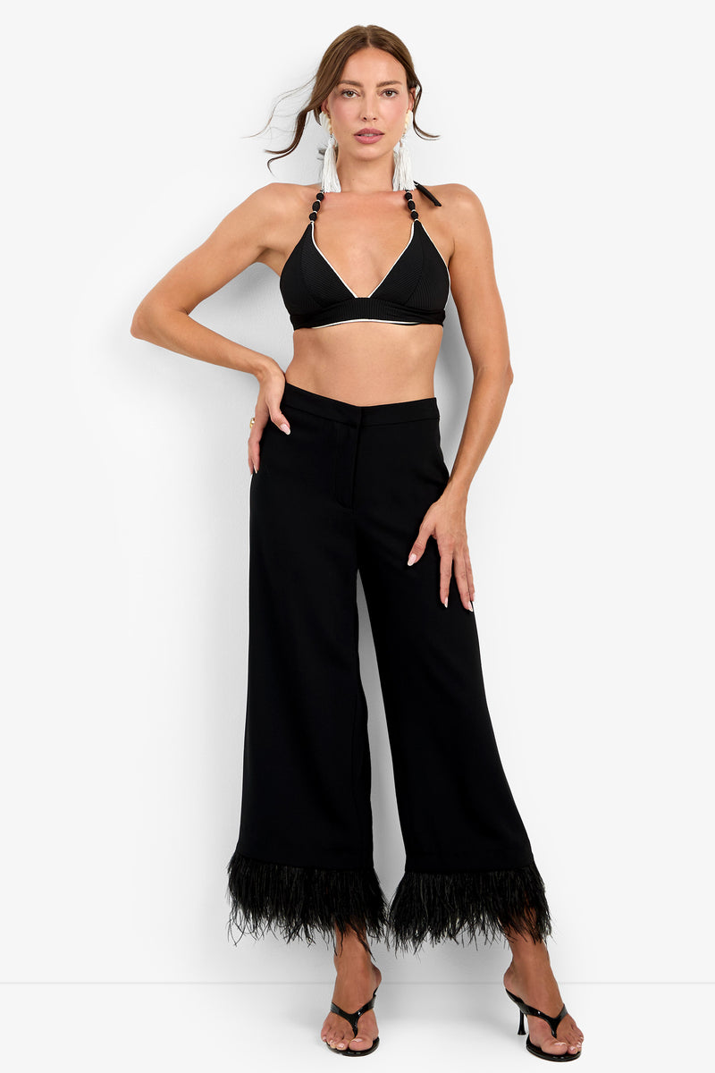 A woman poses against a plain white background wearing the Amarie Pant in black—a wide-leg silhouette featuring feather trim at the hem—styled with a black halter bikini top, black heeled sandals, and statement earrings.