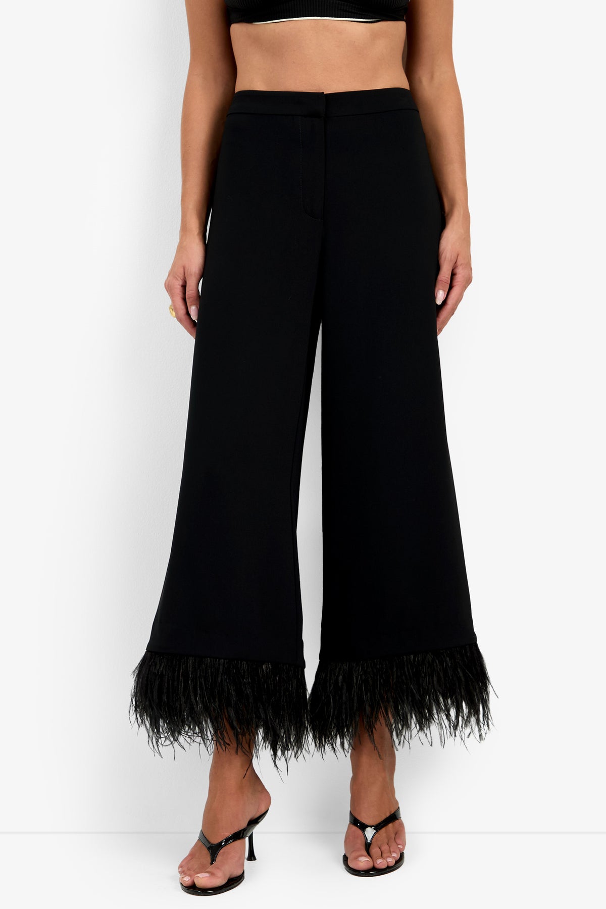Woman wearing the Amarie Pant—a black wide-leg pant with feather trim at the hem—styled with black strappy heels and a black crop top. Only her lower torso and legs are visible against a white background.