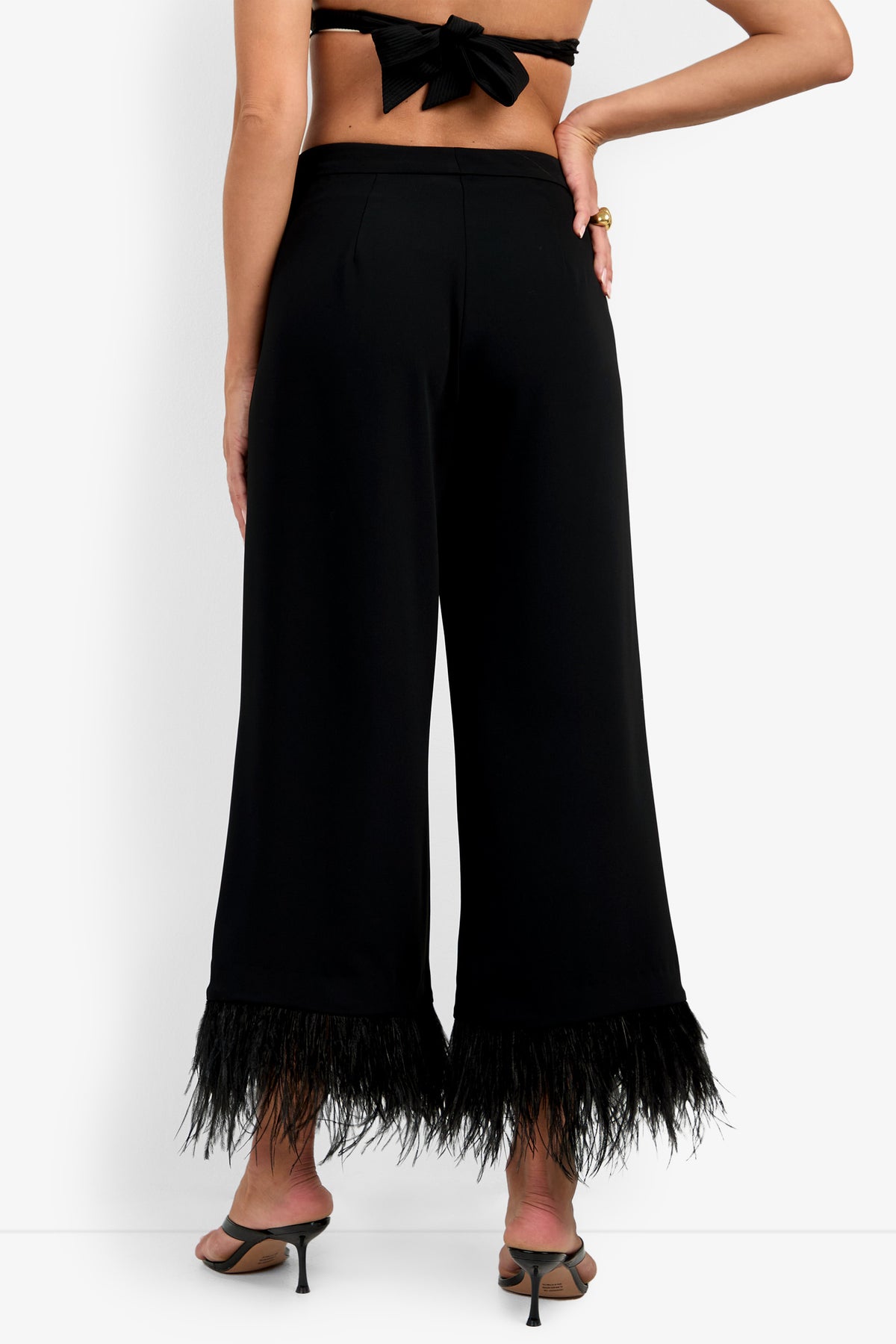 A woman is shown from the back against a plain white background wearing the Amarie Pant—black wide-leg pants featuring feather trim at the hem—paired with black heeled sandals and a tied-back black top.