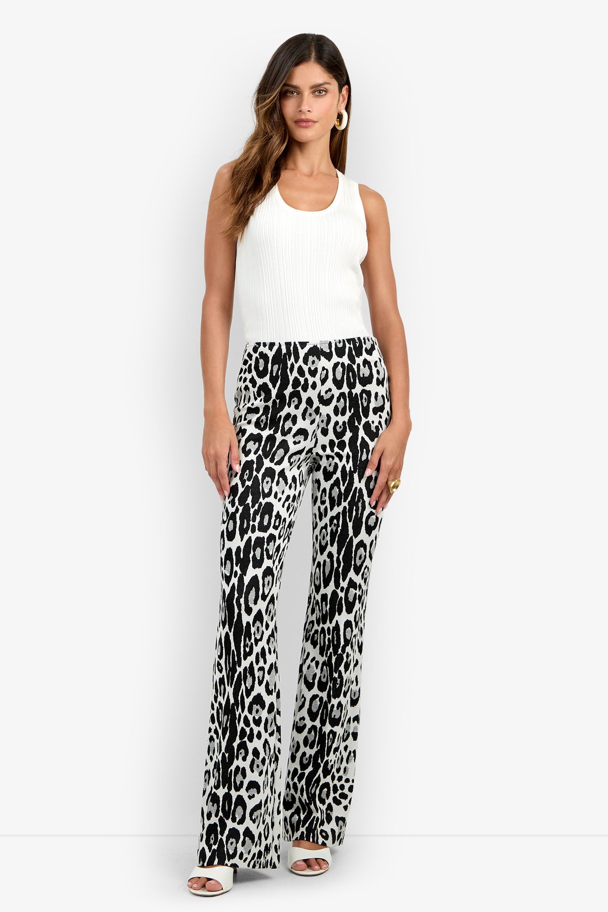 A woman stands against a plain white background, wearing a sleeveless white top paired with the Jacoba Pant—high-waisted, wide-leg black and white animal print pants—along with white heeled sandals, hoop earrings, and a ring.
