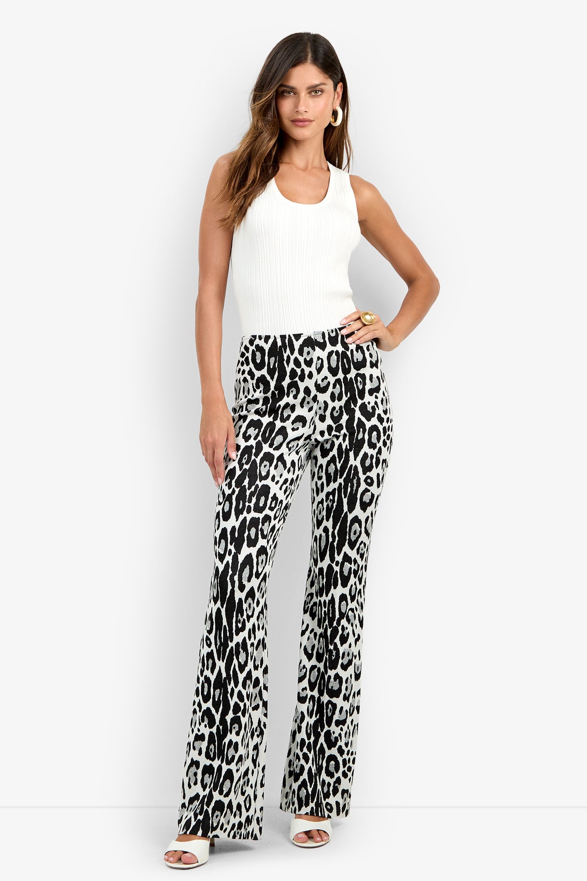 A woman wears the Jacoba Pant in MULTI, featuring a high-waisted, black-and-white leopard print flared silhouette. She pairs it with a sleeveless white top, white sandals, and gold hoop earrings while standing with one hand on her hip.