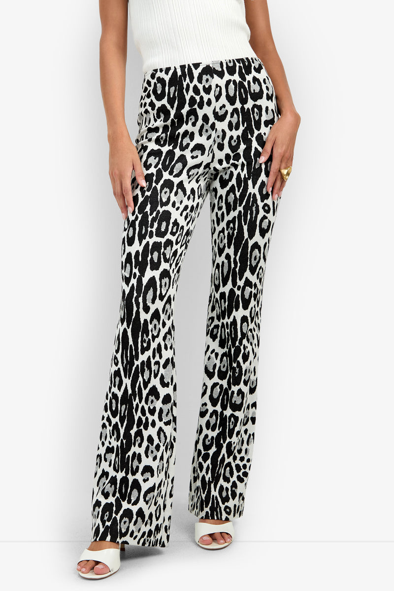 A woman wearing the Jacoba Pant—a pair of high-waisted, wide-leg pants featuring a black and white leopard print—paired with a white sleeveless top, white heels, and a gold ring, stands against a white background.