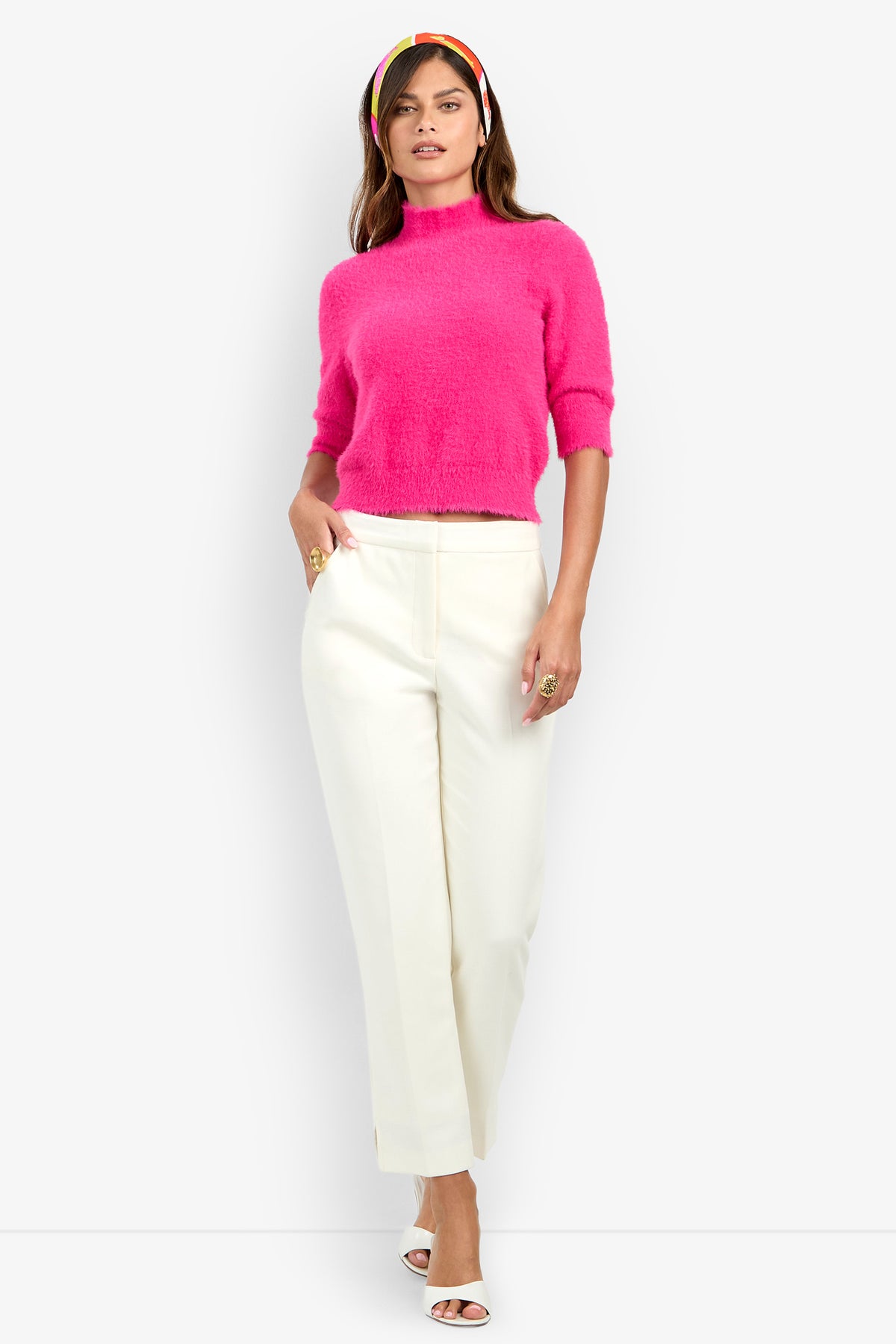 A woman wearing the Arno Pant in white, paired with a bright pink short-sleeve sweater, white open-toe heels, and a colorful headband, stands against a plain white background.