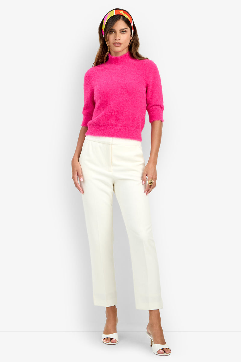 A woman wears the Arno Pant, styled with a bright pink fuzzy sweater, white heeled sandals, and a colorful headband as she stands against a plain light gray background.