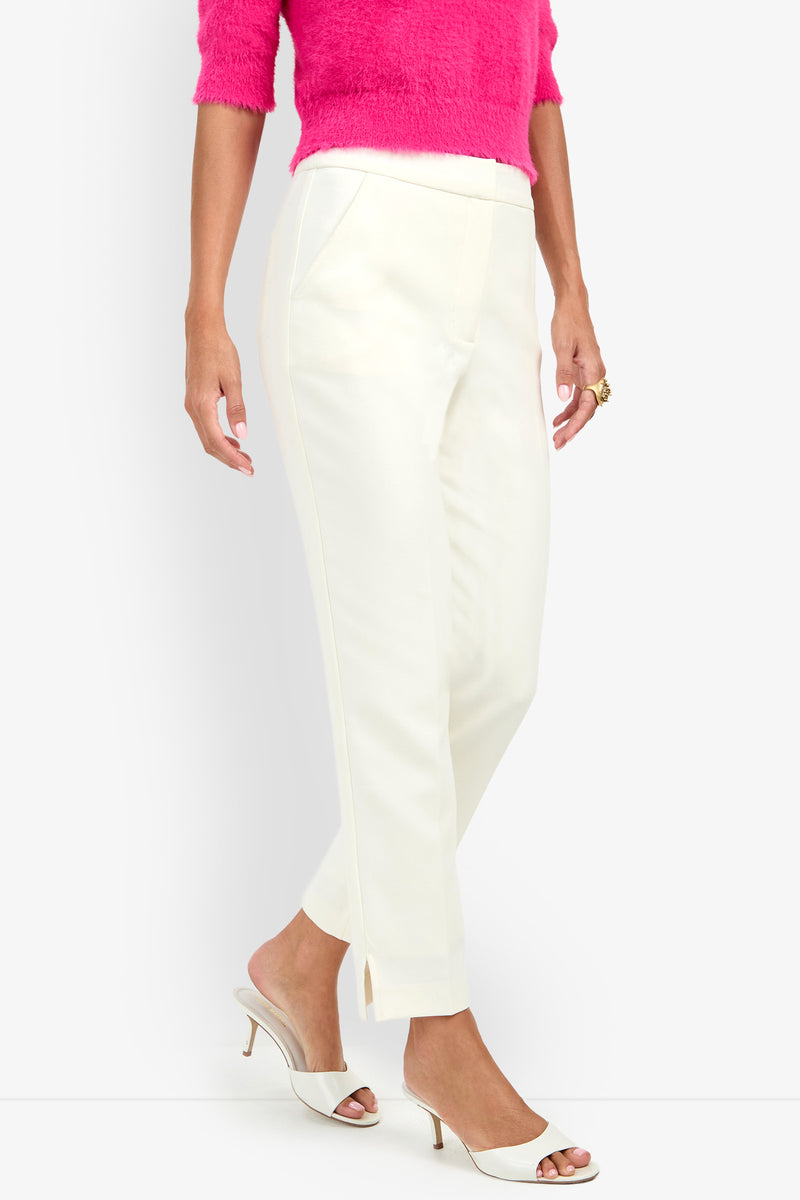 A person wearing the Arno Pant—cream ankle-length pants with small slits at the hem—paired with a bright pink fuzzy sweater and white heeled sandals. Only the lower body is visible.