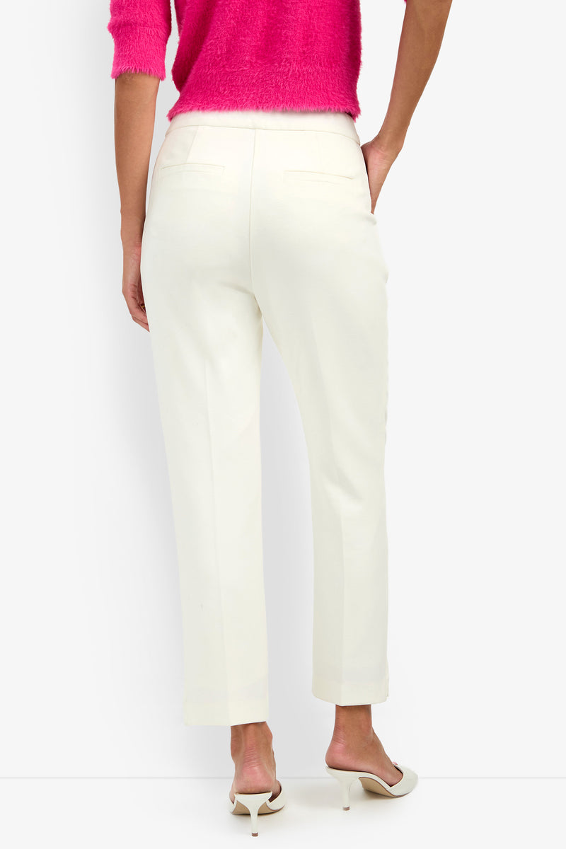 A person wearing the Arno Pant in white—a straight-leg style—with a bright pink top, standing with one hand in their pocket. They are also wearing white heeled sandals.