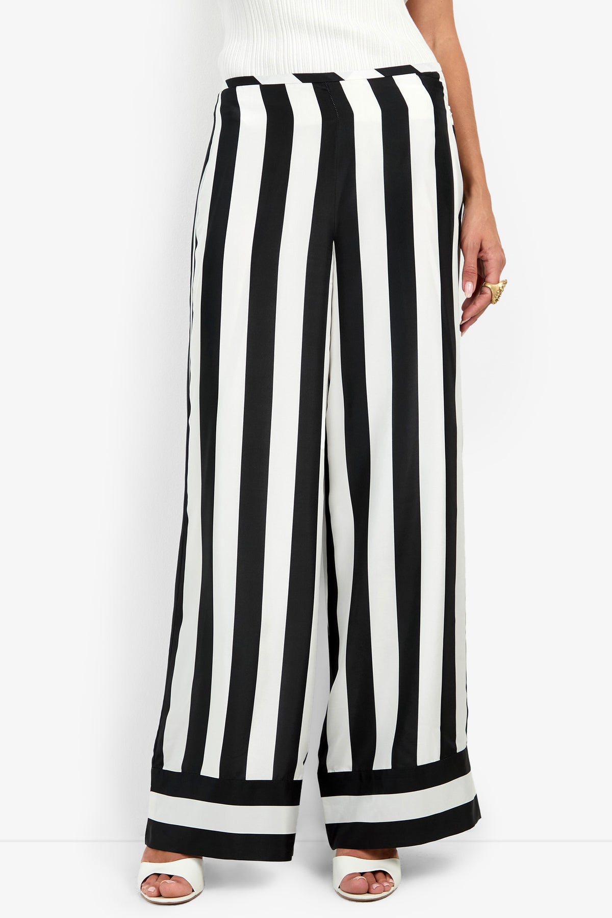 A person wears the Khiara Pant, featuring wide-leg, high-waisted bold vertical black and white stripes, paired with white open-toe heels and a white top.