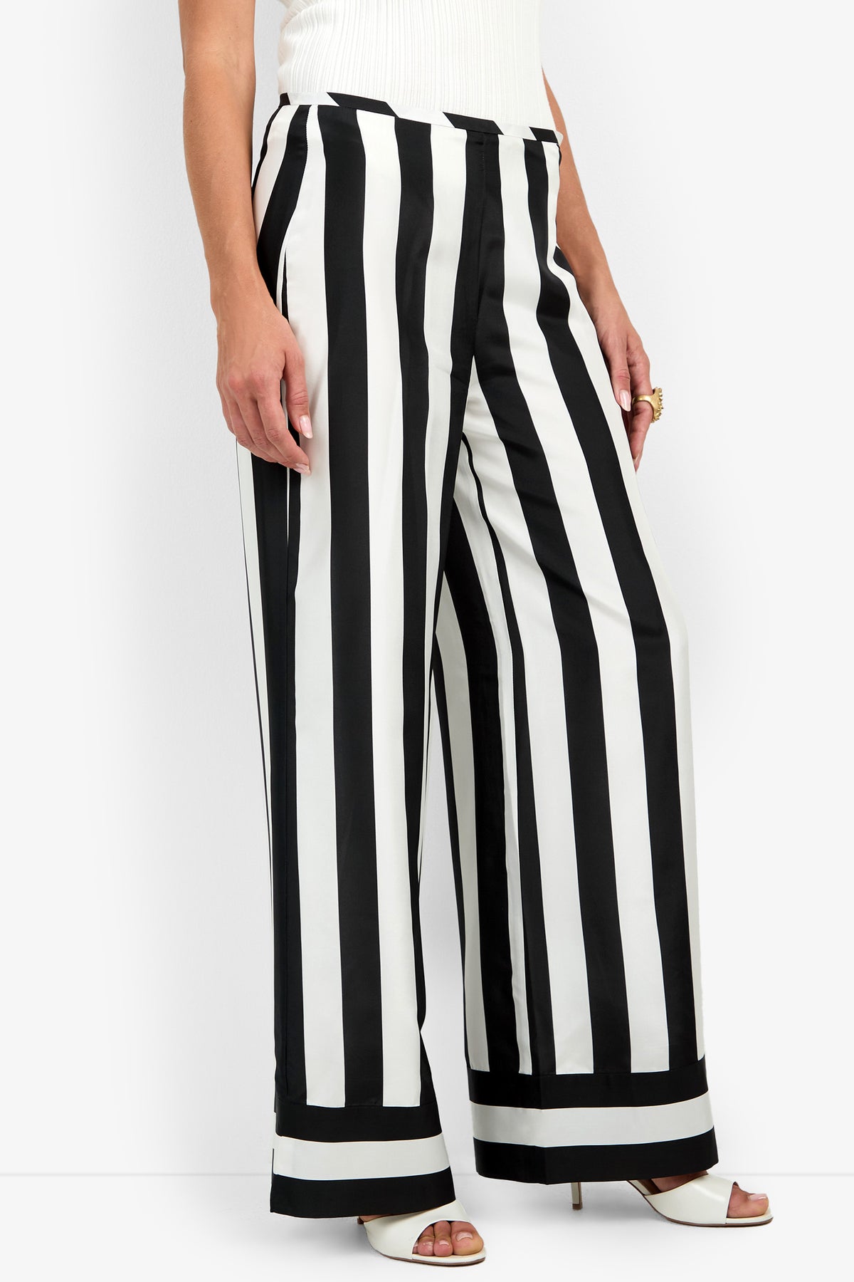 A person wearing the Khiara Pant—a pair of high-waisted, wide-leg black and white vertical striped pants—paired with white heeled sandals and a white top. Only the lower half of the body is visible.