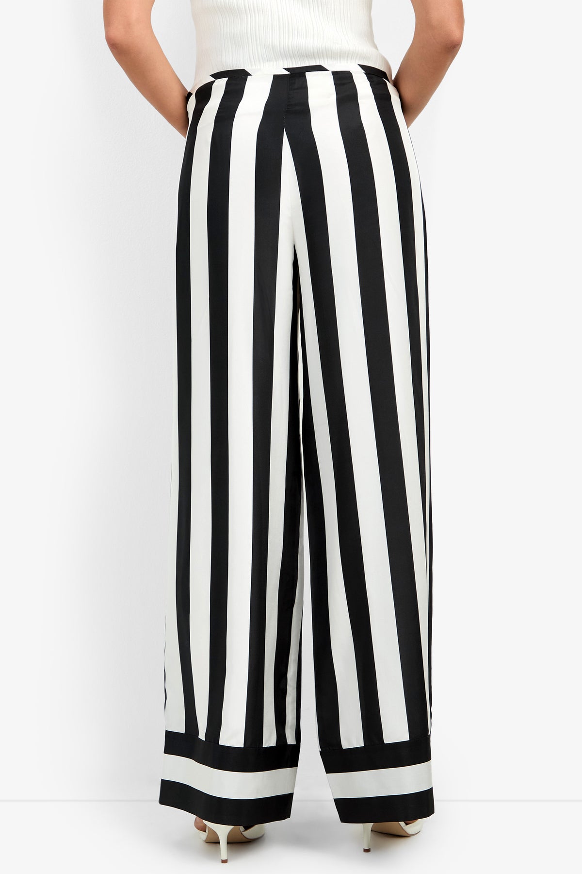 A person seen from the back wearing the Khiara Pant—high-waisted, wide-leg trousers featuring bold vertical black and white stripes—paired with light heels and a tucked-in white top.