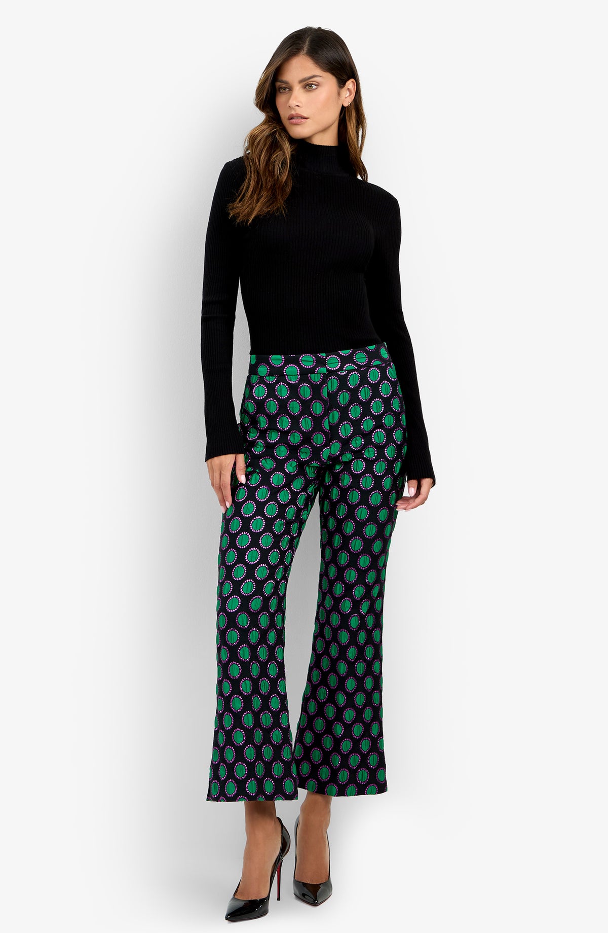 A woman stands against a plain white background wearing a black turtleneck sweater, black high heels, and the Allisan Pant—wide-leg pants featuring a green and purple pattern.