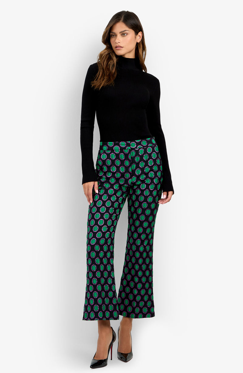 A woman stands against a plain white background wearing a black turtleneck sweater, black high heels, and the Allisan Pant—wide-leg pants featuring a green and purple pattern.