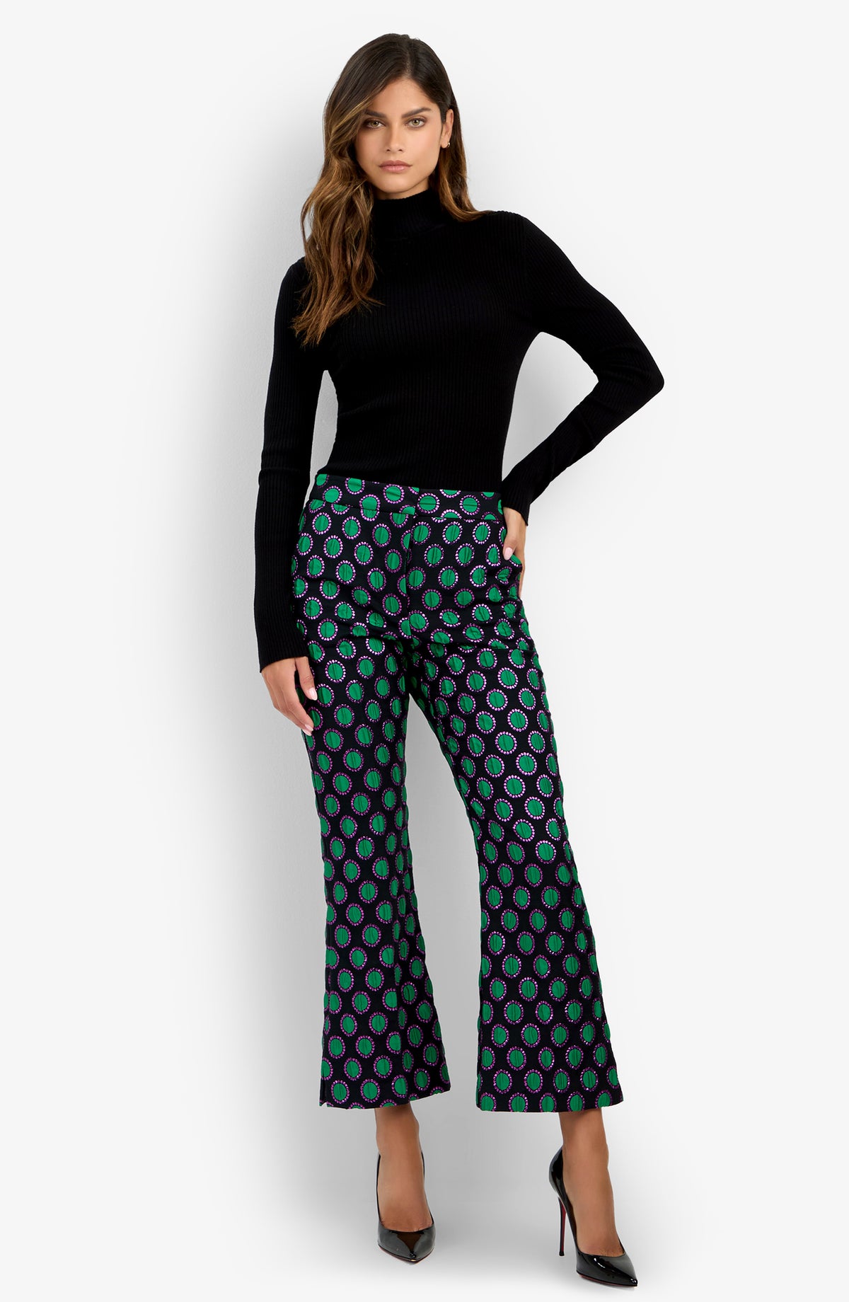 A woman wearing a black turtleneck sweater, the Allisan Pant—featuring a blue and green pattern with a flared silhouette—and black high-heeled shoes, standing against a plain white background.
