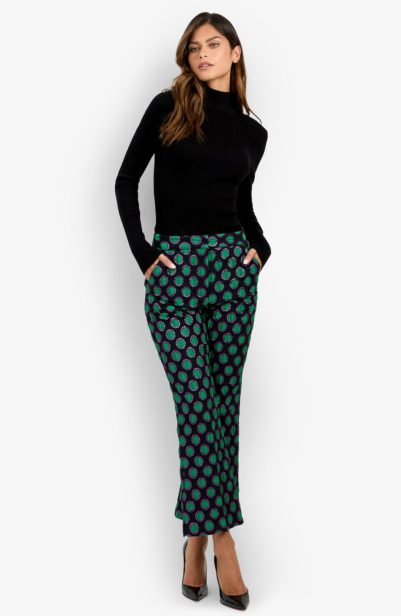 A woman stands with her hands in her pockets, wearing a black long-sleeve top, the patterned green and purple Allisan Pant, and black high heels against a plain white background.