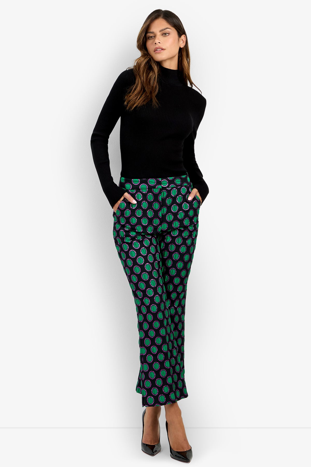 A woman stands against a plain white background, hands in pockets, wearing the Allisan Pant—a pair of green patterned pants—paired with a black long-sleeve turtleneck and black heels.