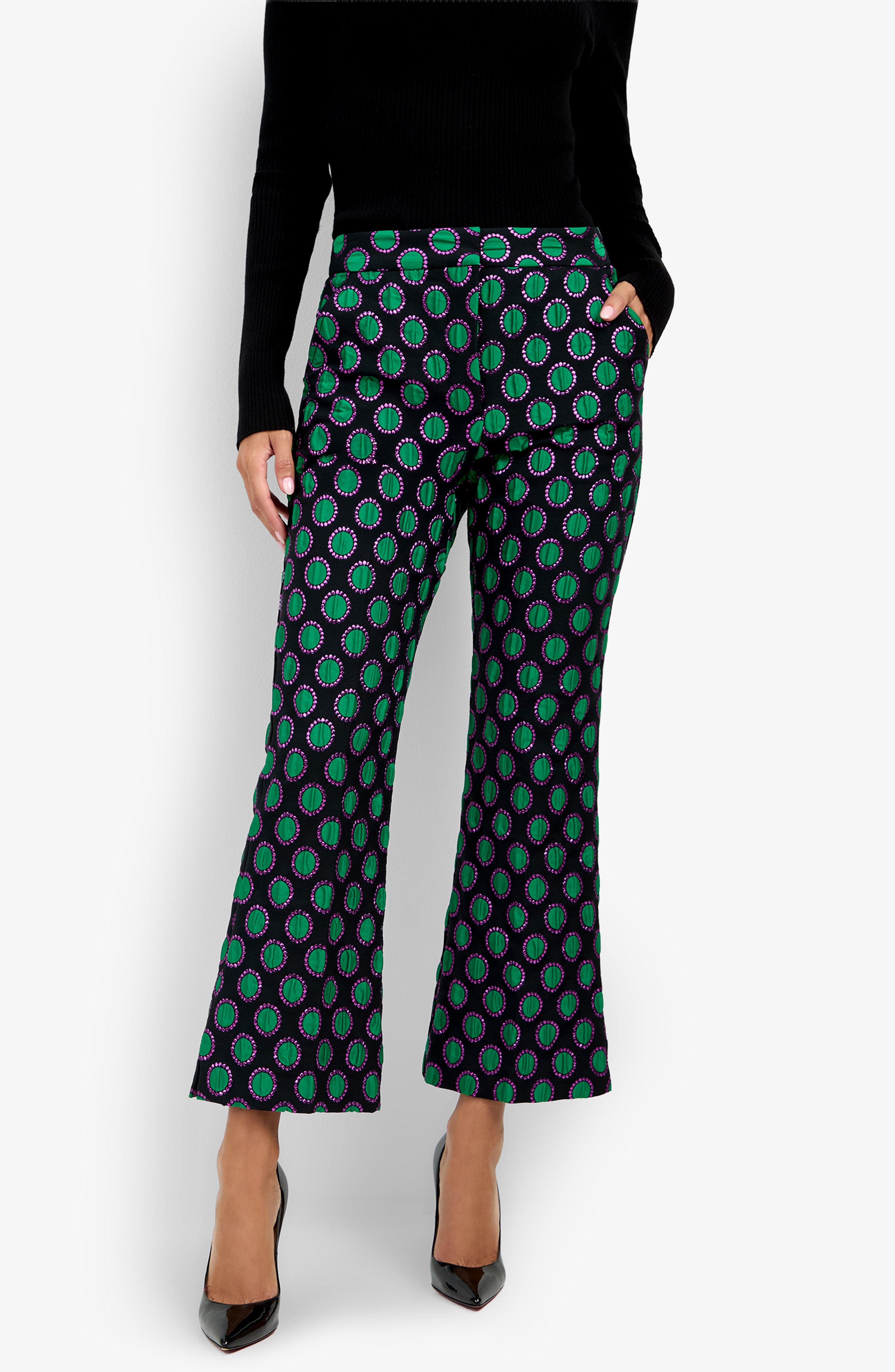 Model wearing the Allisan Pant—flared, patterned pants with green and purple circles—paired with a black long-sleeve top and black pointed-toe heels, posing with one hand in a pocket against a plain white background.