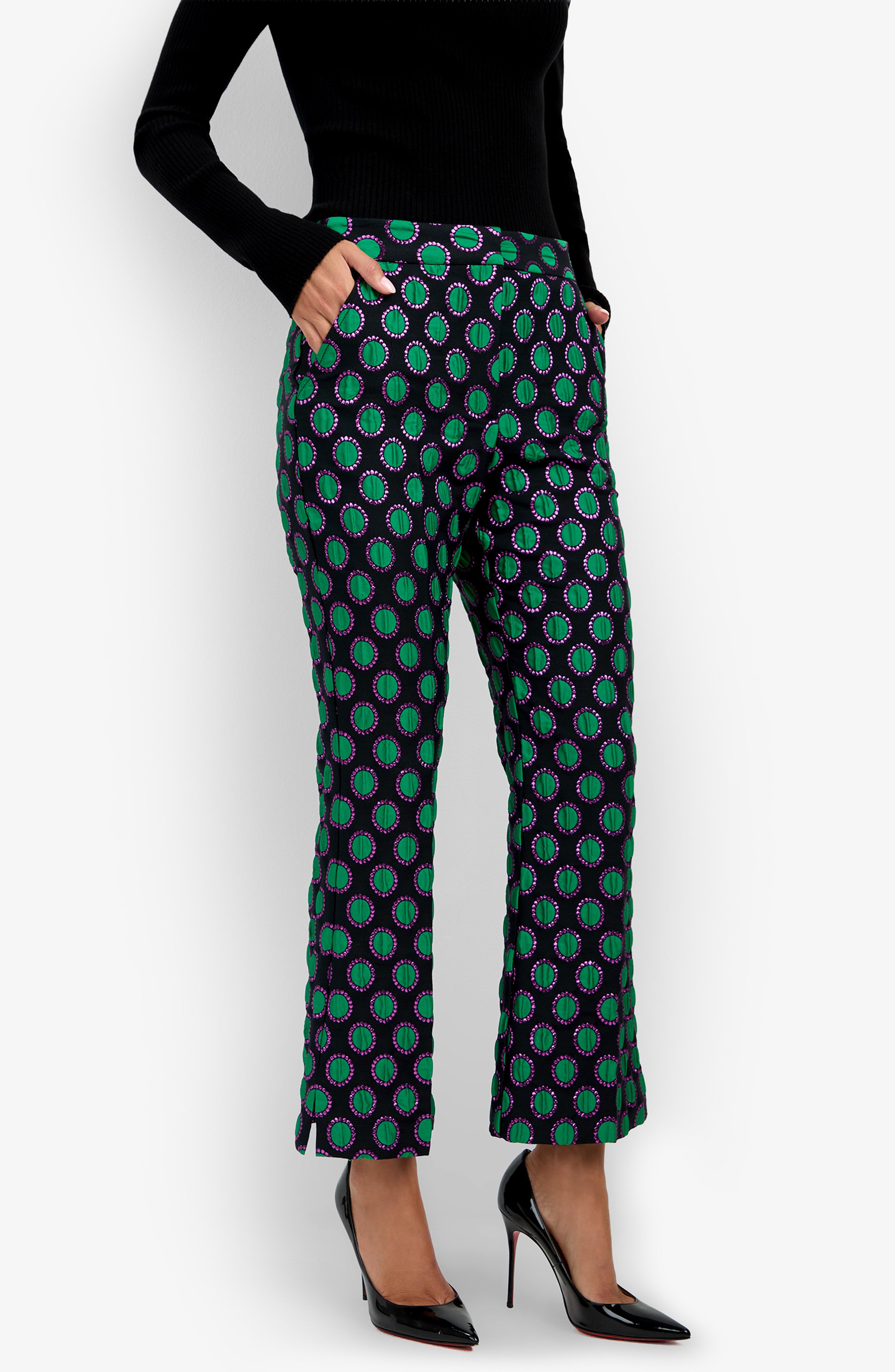 A woman in a black turtleneck wears the Allisan Pant—patterned in green, purple, and blue—with her hands in her pockets and paired with black high-heeled shoes, standing against a white background.