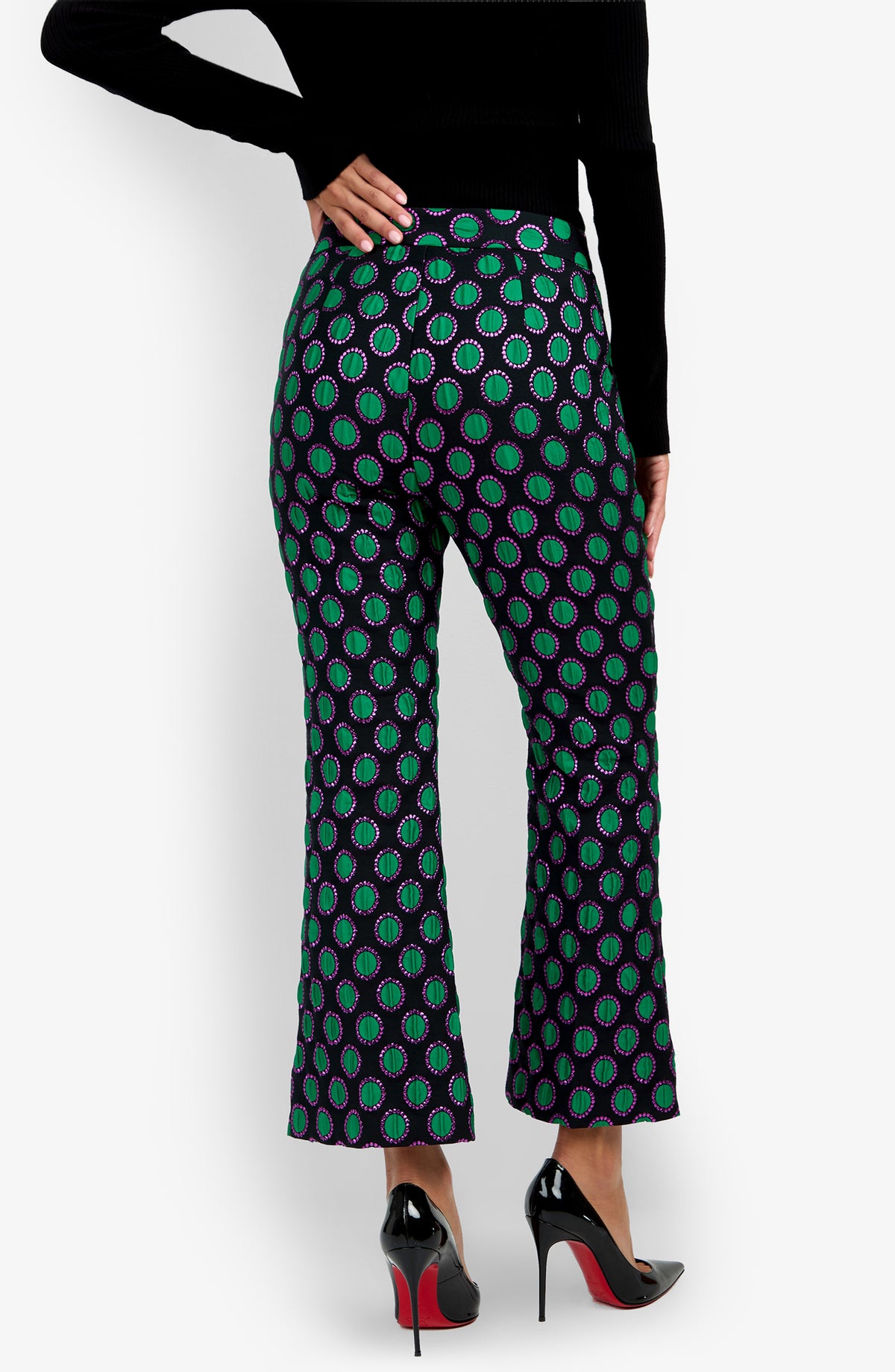 Person wearing the Allisan Pant, a pair of high-waisted, flared pants with a green and purple circular pattern, styled with black high heels and a black long-sleeve top, seen from the back.