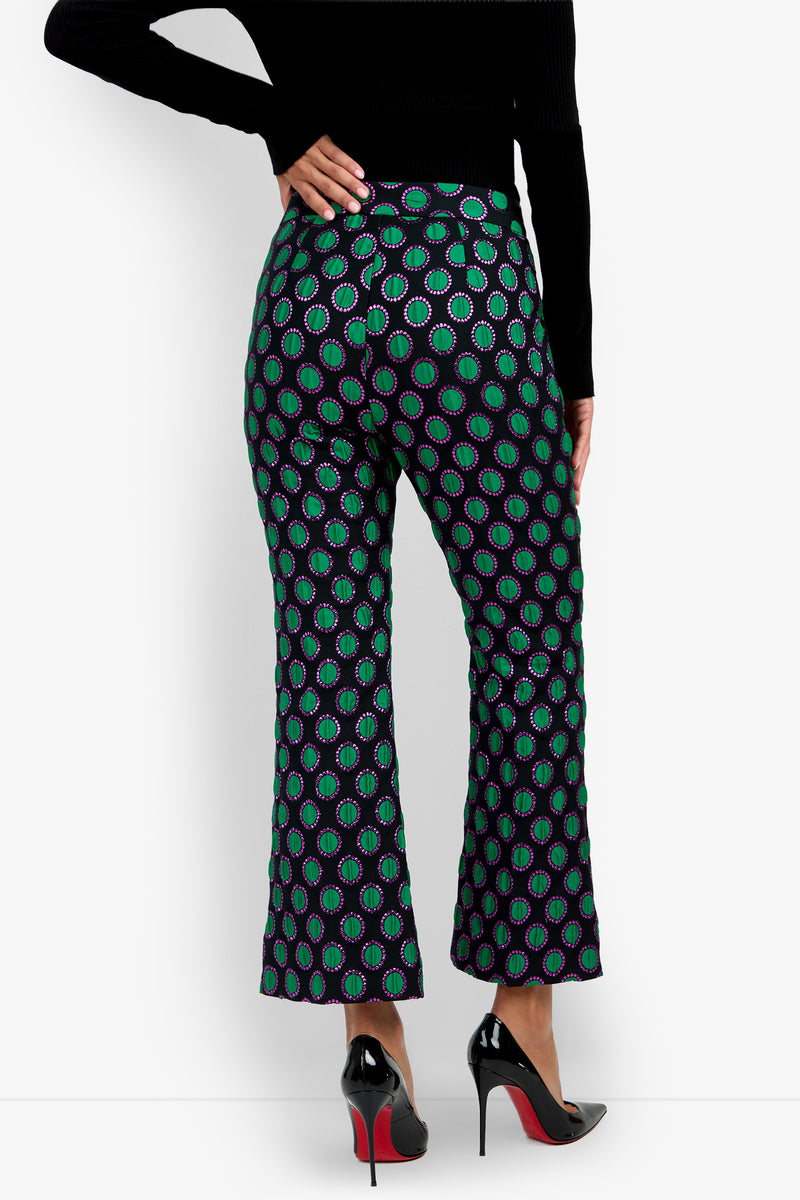 A person is seen from the back wearing the Allisan Pant, which features a vibrant green and purple geometric pattern and a flared silhouette, paired with black high heels and a black long-sleeve top.