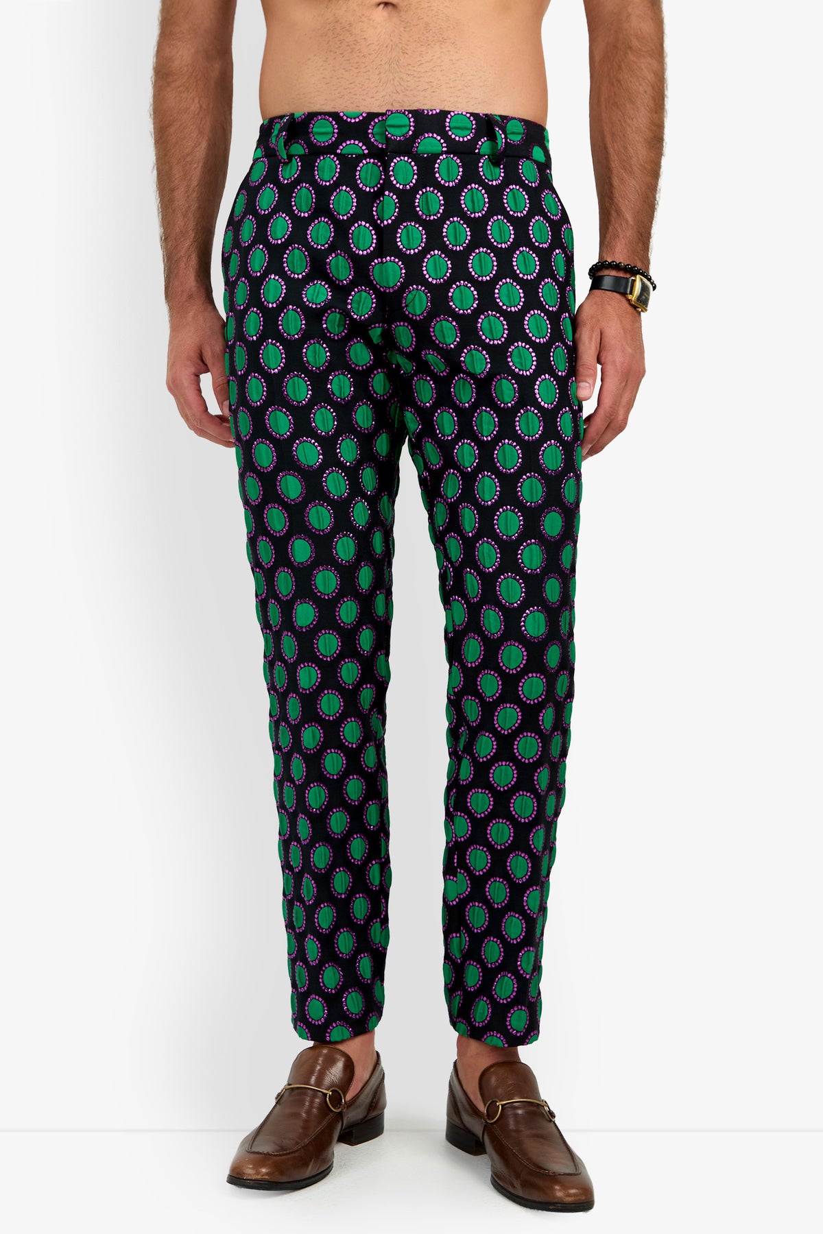 A man wears Clyde Slim Trouser featuring a bold pattern of green and purple circles on a dark background, paired with brown loafers and a watch. His torso is not shown.