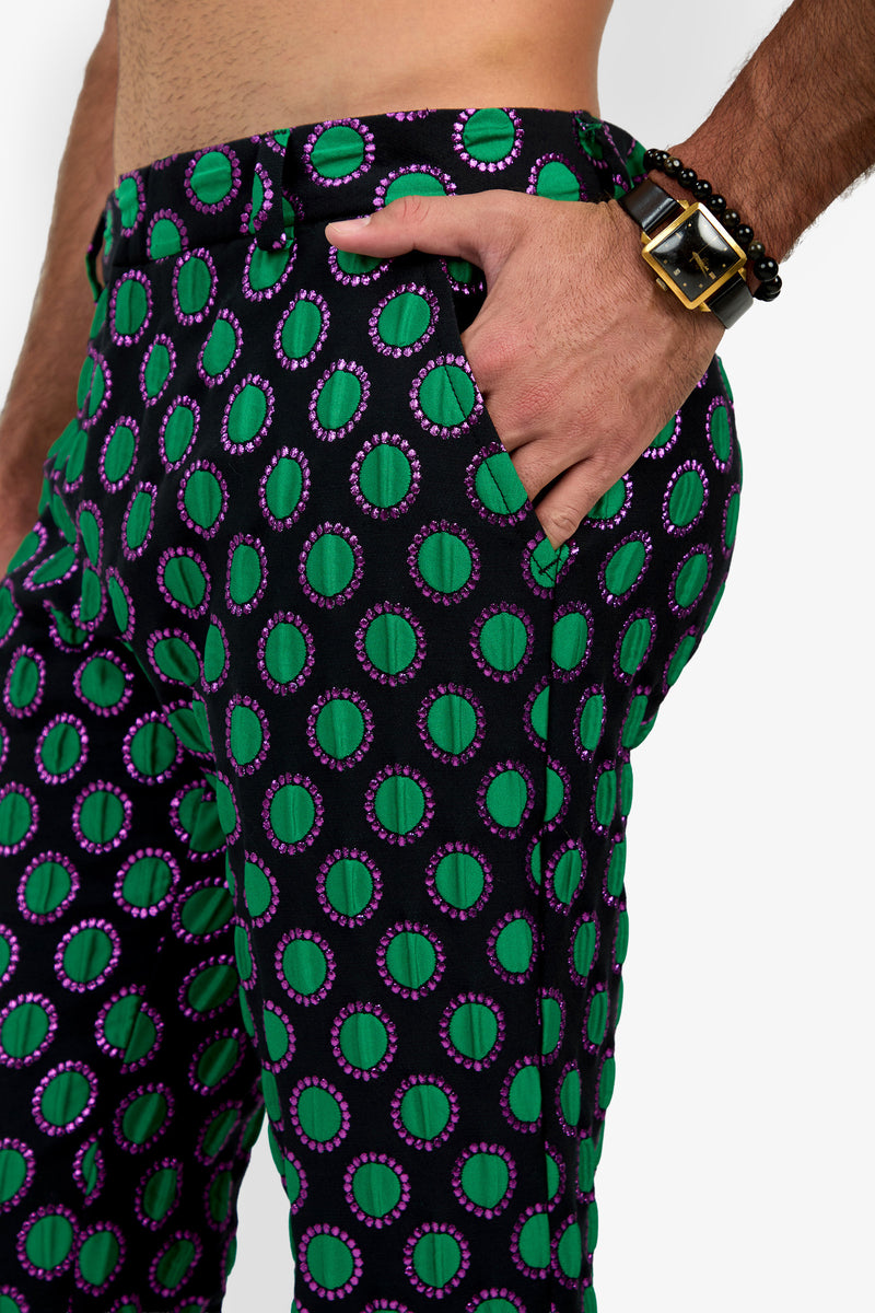 A person wearing the Clyde Slim Trouser, featuring black fabric with green and purple circular patterns, has their right hand in their pocket, revealing a black and gold watch along with beaded bracelets.