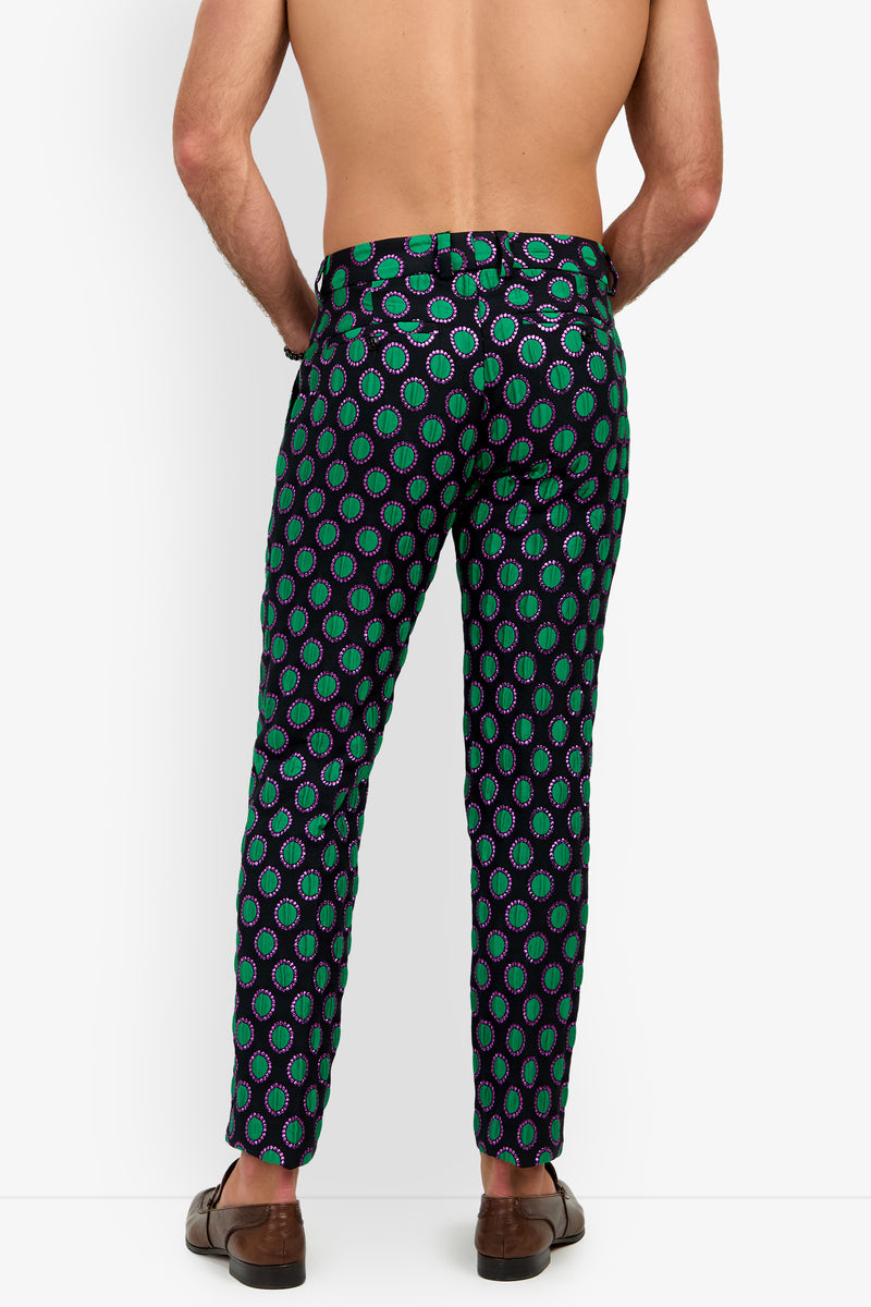 A shirtless person standing facing away from the camera against a plain white background, wearing Clyde Slim Trouser featuring a dark pattern with green and purple circles, paired with brown loafers.