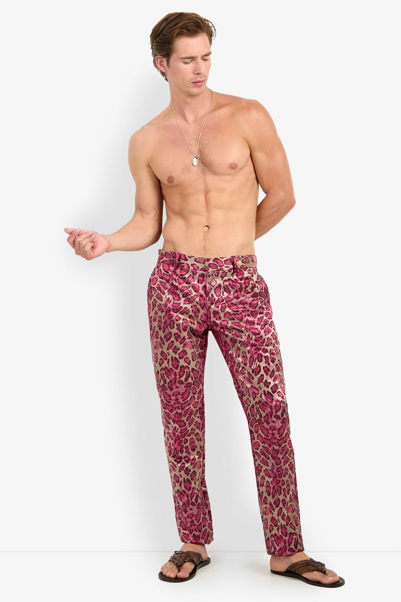 A shirtless man stands against a plain white background wearing the CLYDE SLIM TROUSER in pink leopard print, paired with sandals and a necklace. One hand is in his pocket while the other is slightly raised.