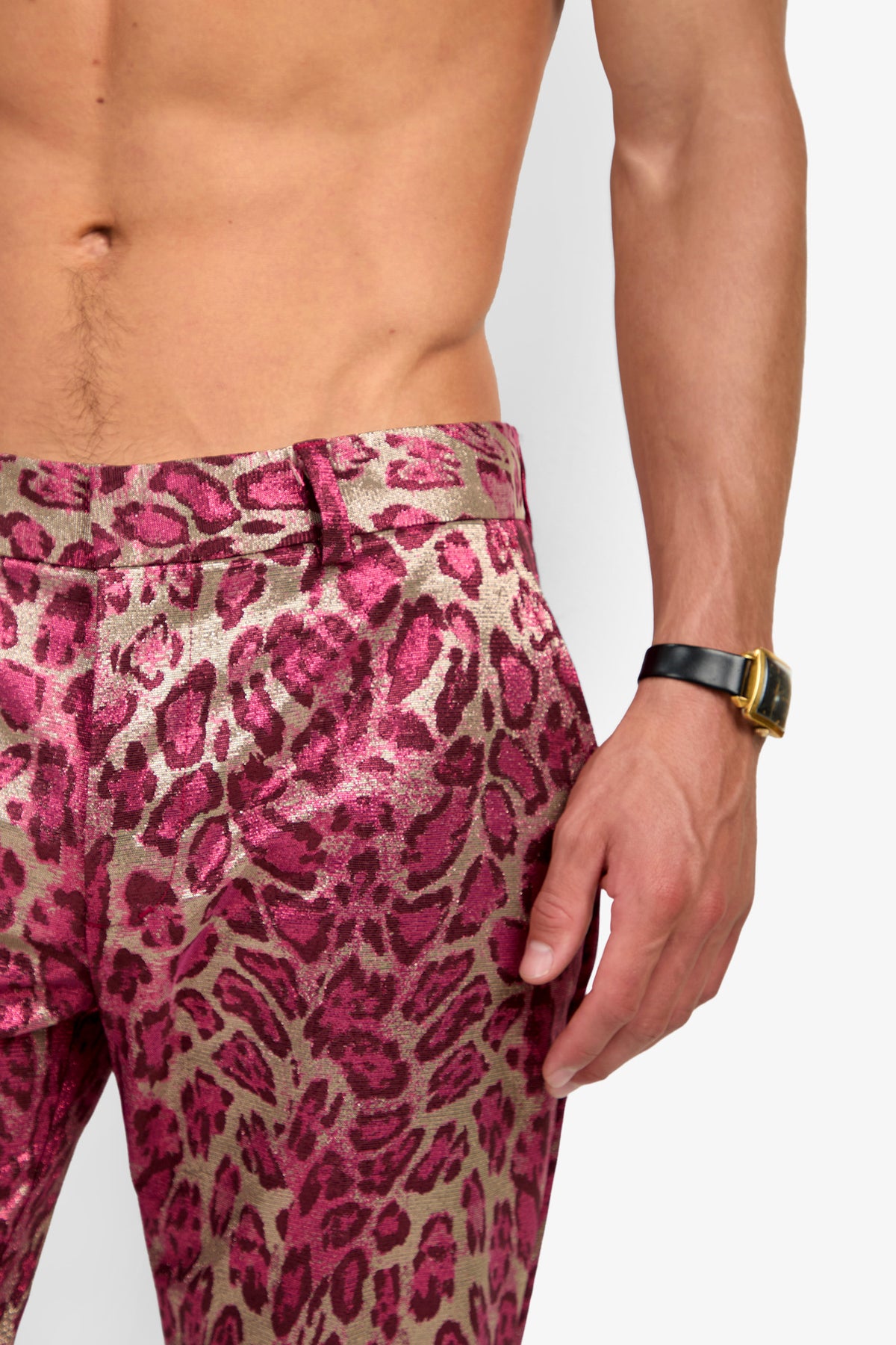 A shirtless person wearing the CLYDE SLIM TROUSER—shiny pink and gold leopard print pants—and a black wristwatch, with their right hand resting by their side.
