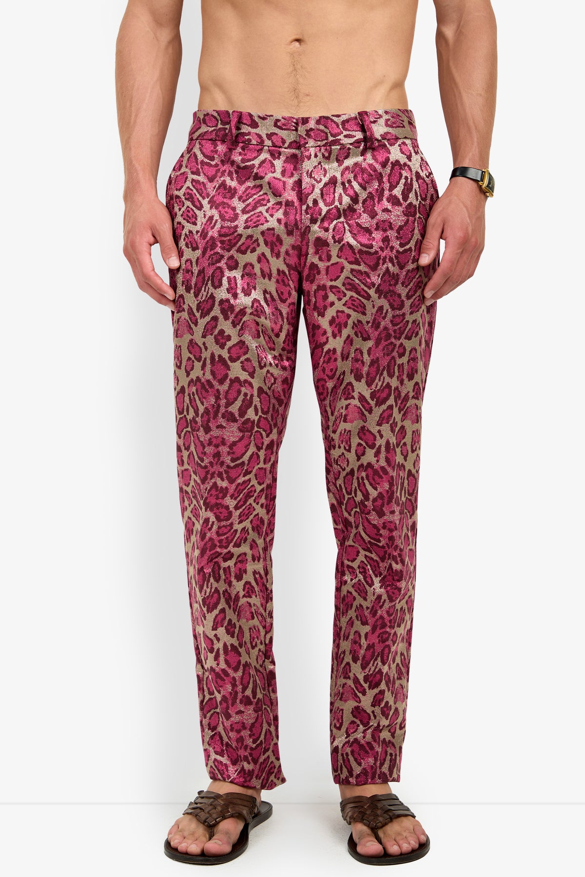 A shirtless man wears the CLYDE SLIM TROUSER in pink leopard print, paired with brown sandals and a wristwatch, standing against a plain white background.