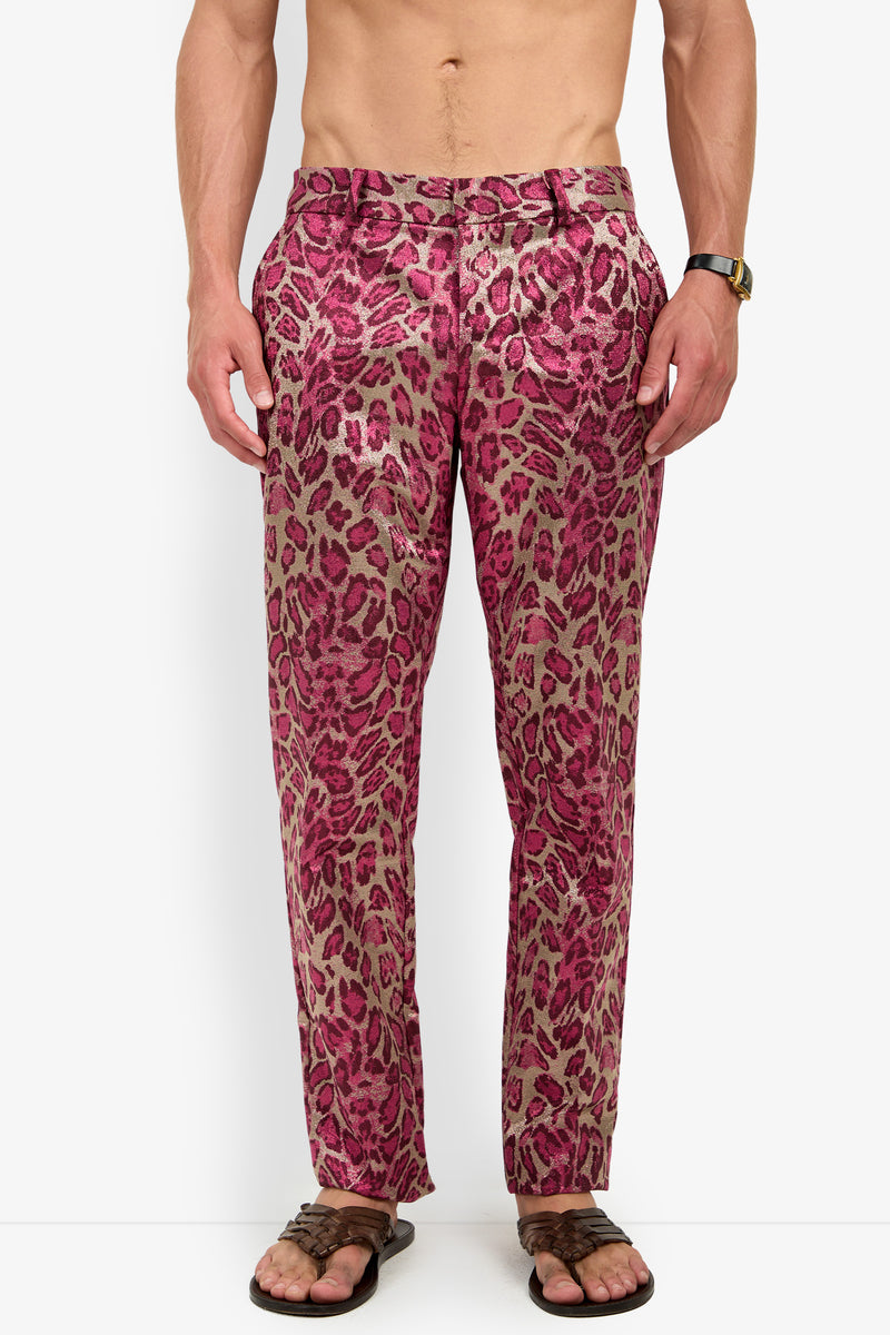 A shirtless man wears the CLYDE SLIM TROUSER in pink leopard print, paired with brown sandals and a wristwatch, standing against a plain white background.