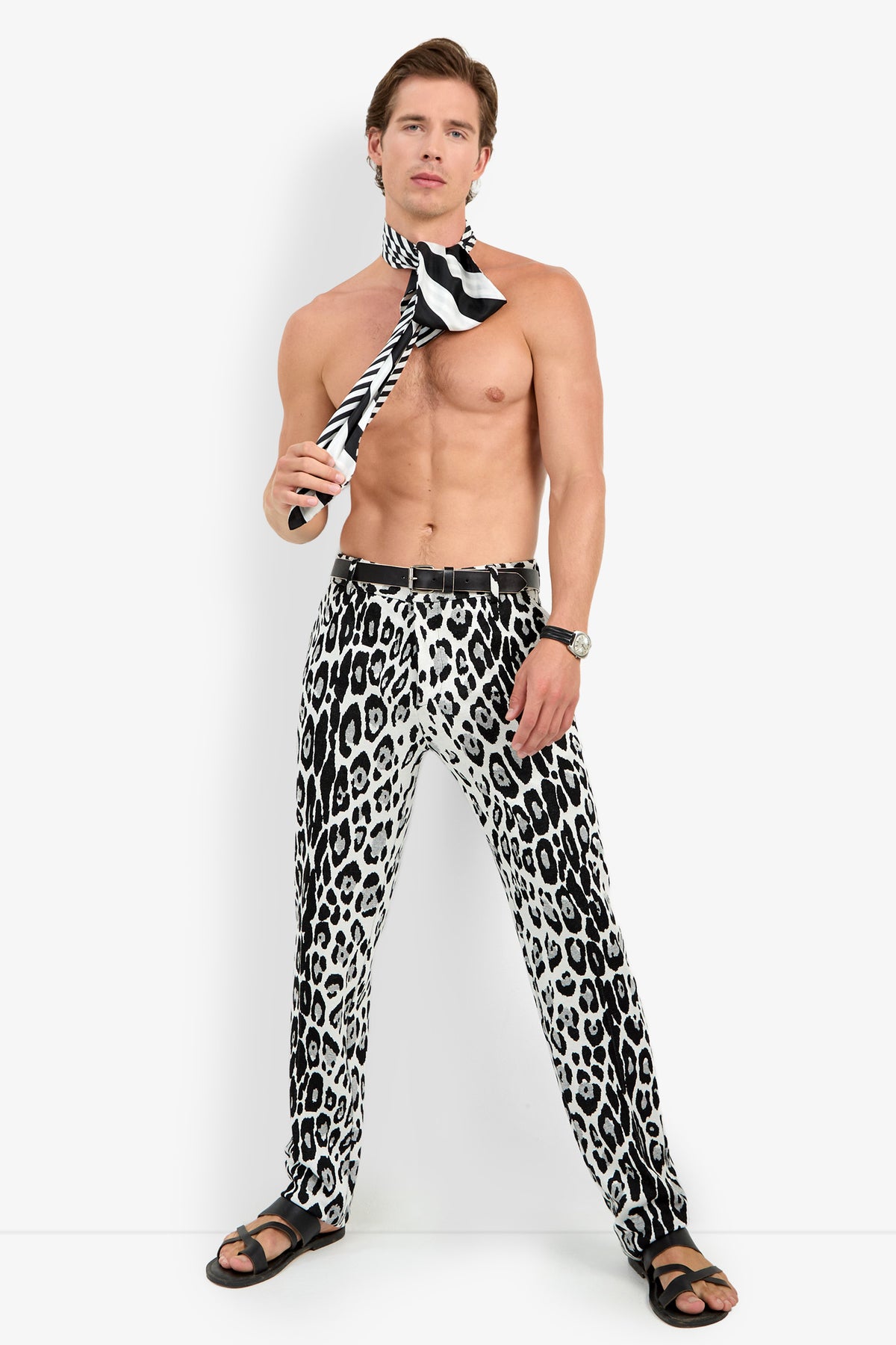 Shirtless man standing, wearing the Clyde Slim Trouser in black-and-white animal print, a matching striped scarf around his neck, sandals, and a watch, posing against a plain white background.