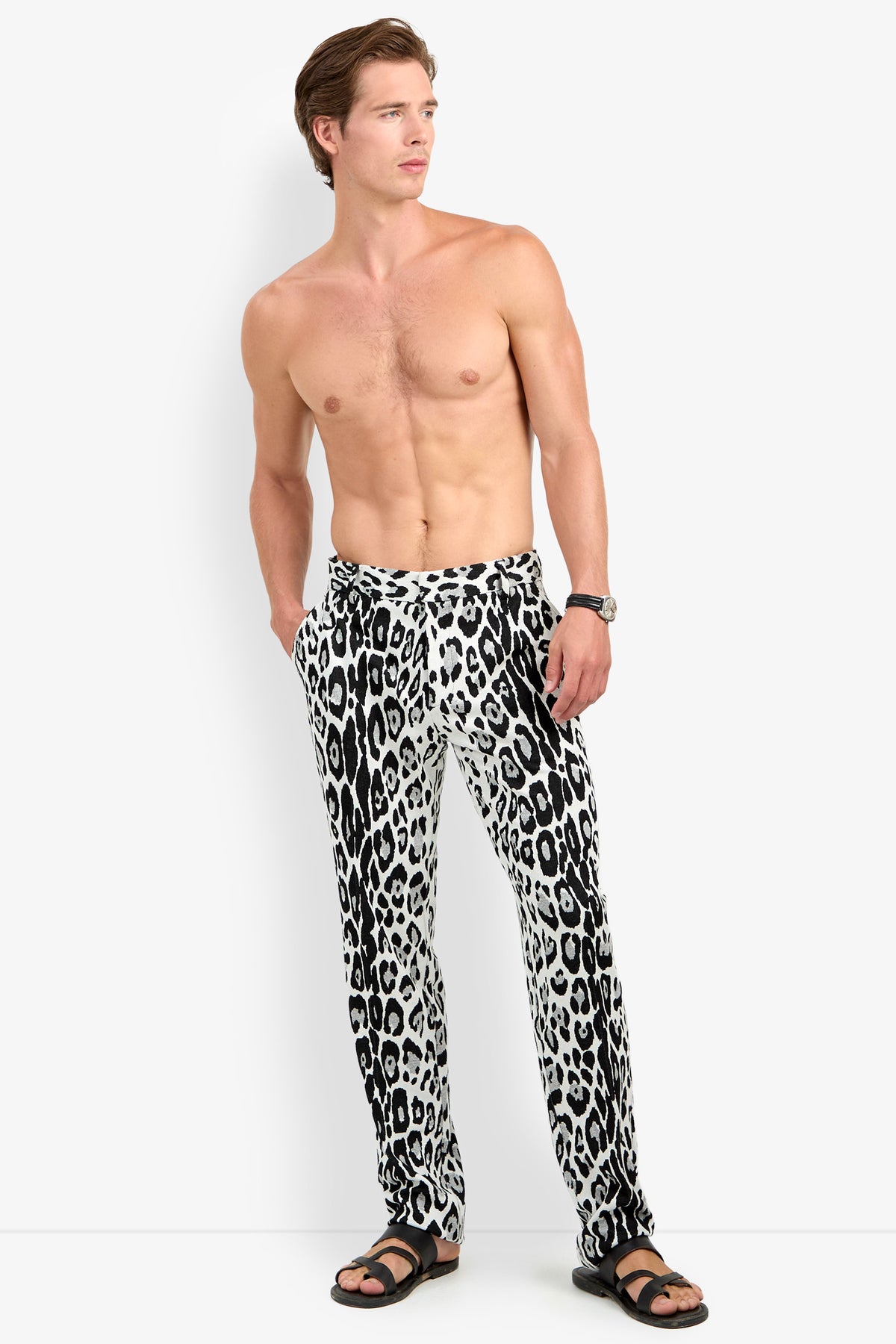 A shirtless man wears the Clyde Slim Trouser in a black-and-white leopard print, paired with black sandals and a wristwatch. He stands with his hands in his pockets, looking to the side against a plain background.