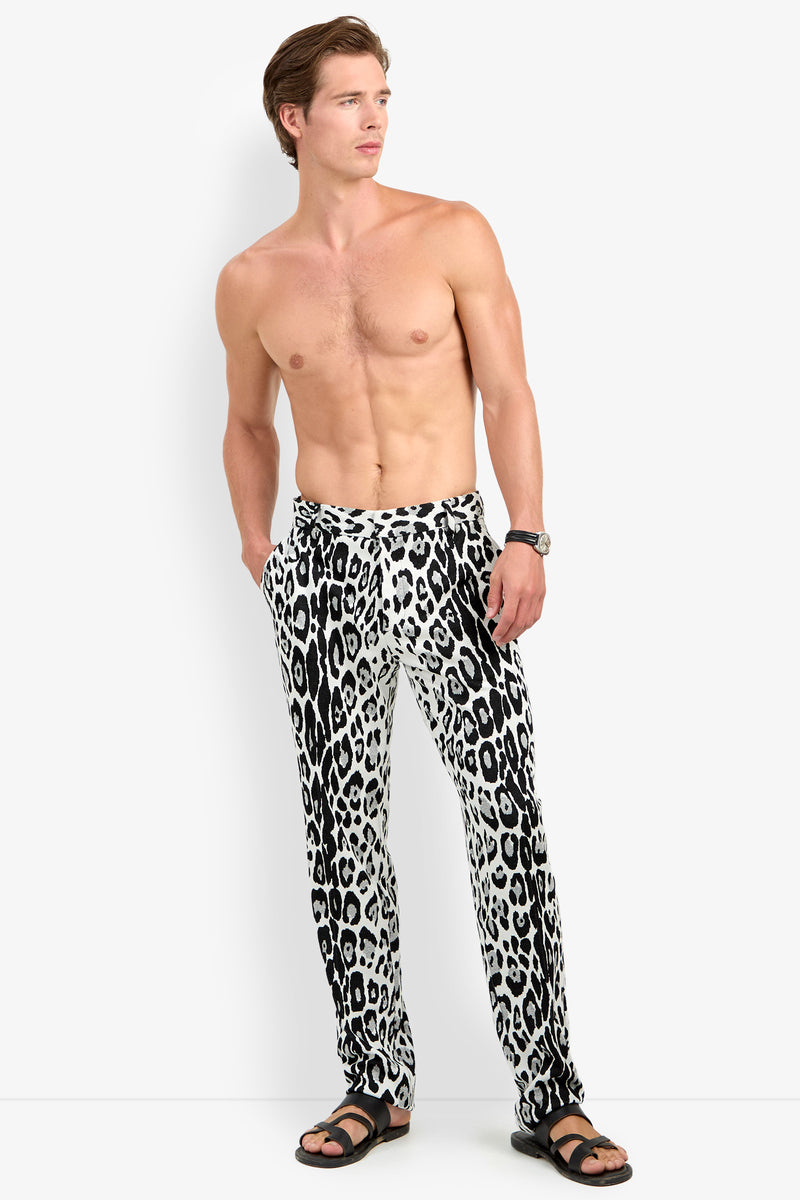 A shirtless man wears the Clyde Slim Trouser in a black-and-white leopard print, paired with black sandals and a wristwatch. He stands with his hands in his pockets, looking to the side against a plain background.