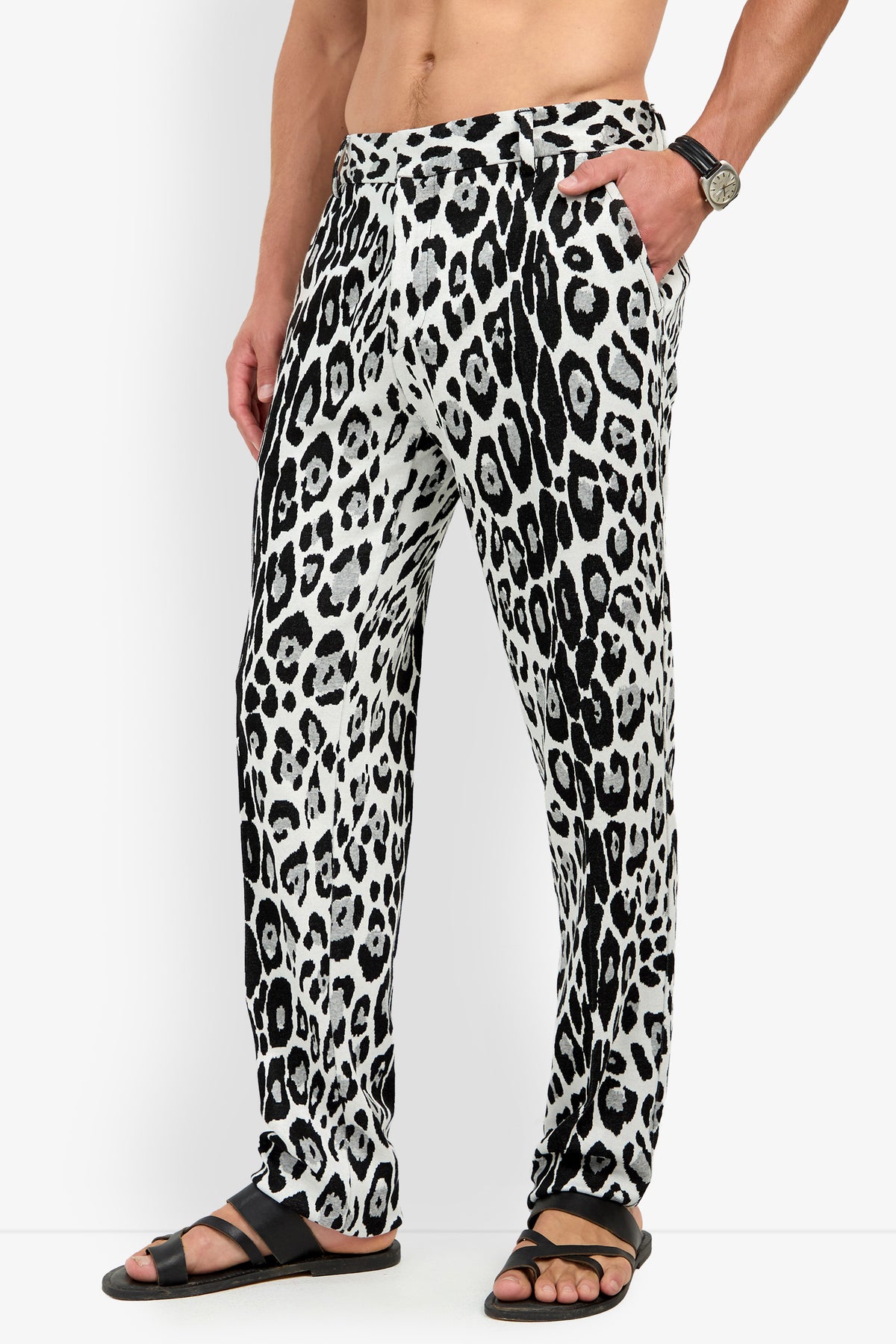 A person stands against a plain background with one hand in their pocket, wearing the Clyde Slim Trouser in a black and white leopard print, paired with black sandals and a wristwatch.
