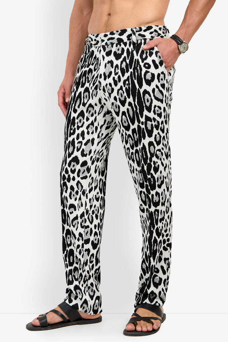 A person stands against a plain background with one hand in their pocket, wearing the Clyde Slim Trouser in a black and white leopard print, paired with black sandals and a wristwatch.