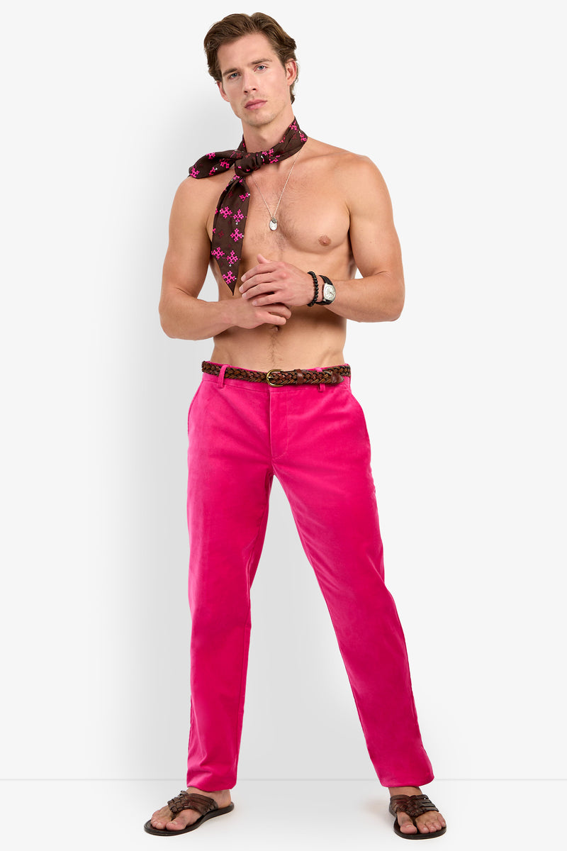 A shirtless man wears the Clyde Slim Trouser in bright pink, paired with a brown patterned scarf tied around his neck, a watch, a necklace, and flip-flops. He stands in a relaxed pose against a plain white background.