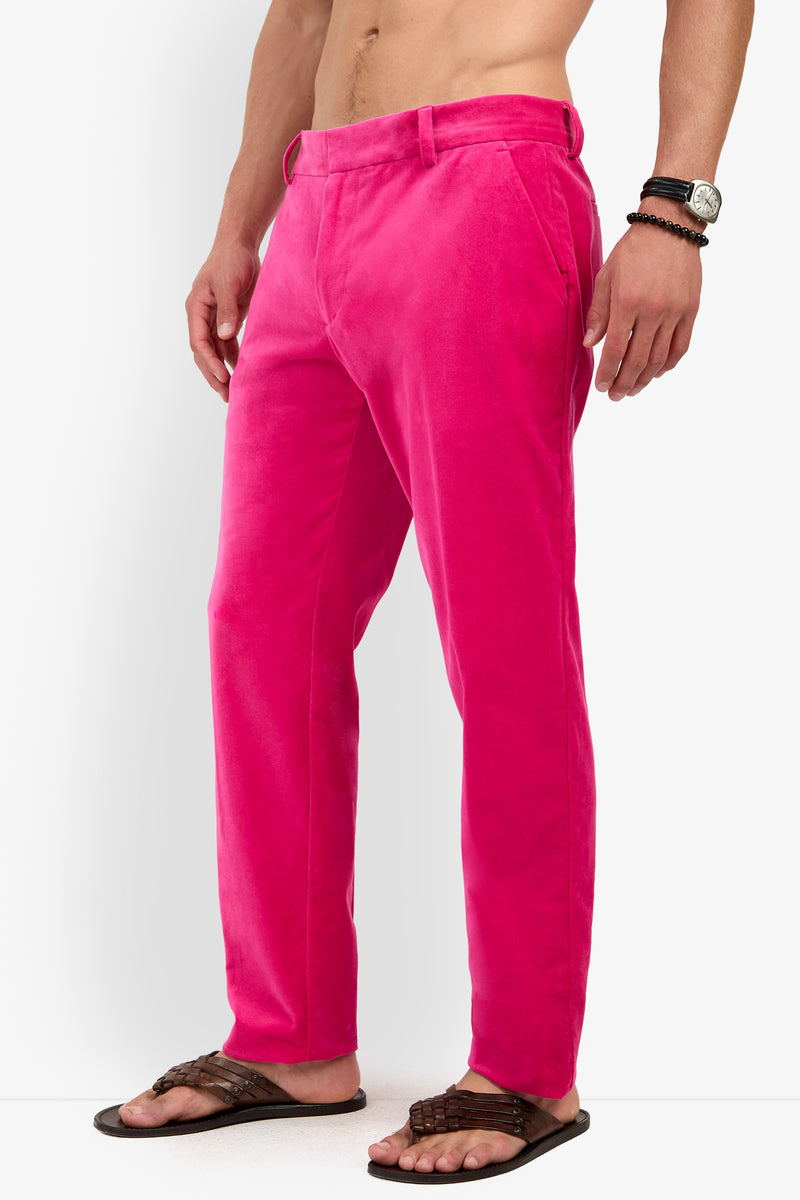 A person wearing the Clyde Slim Trouser in bright pink, paired with dark brown sandals, a black watch, and bracelets, standing against a white background.
