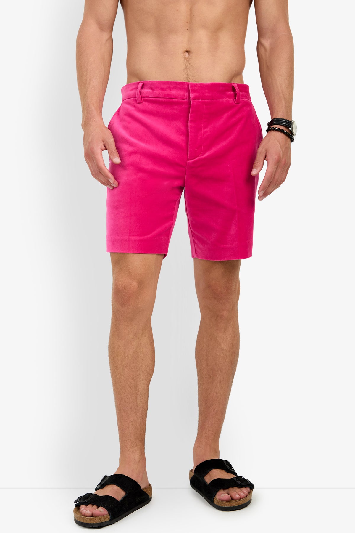 A shirtless man models the LAWRENCE SHORT in bright pink, paired with black sandals and wrist accessories, standing against a plain white background.