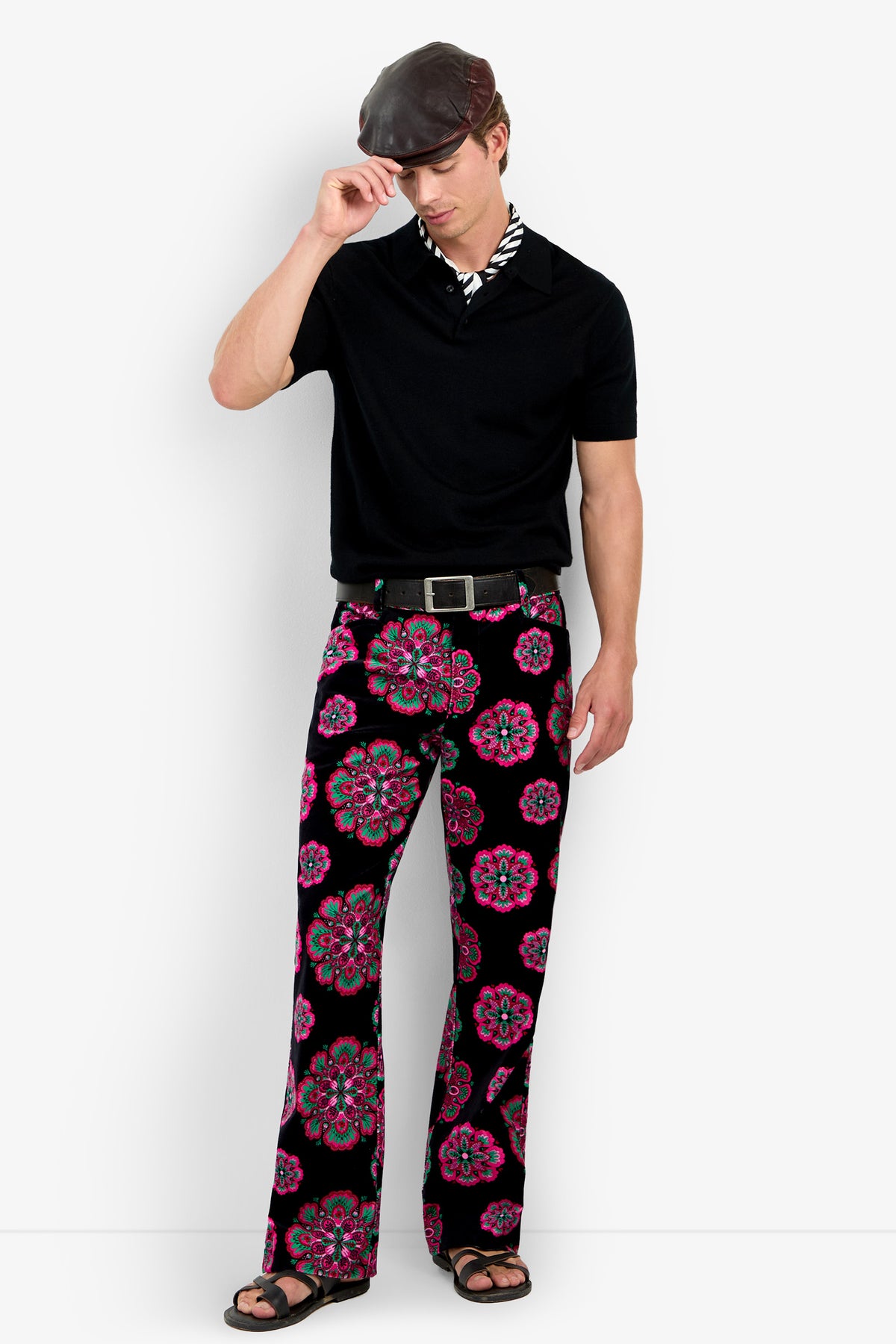 A man wearing a black shirt, the Alejandro Bootleg Trouser with colorful floral patterns, sandals, and a flat cap stands against a white background, touching the brim of his hat with one hand.