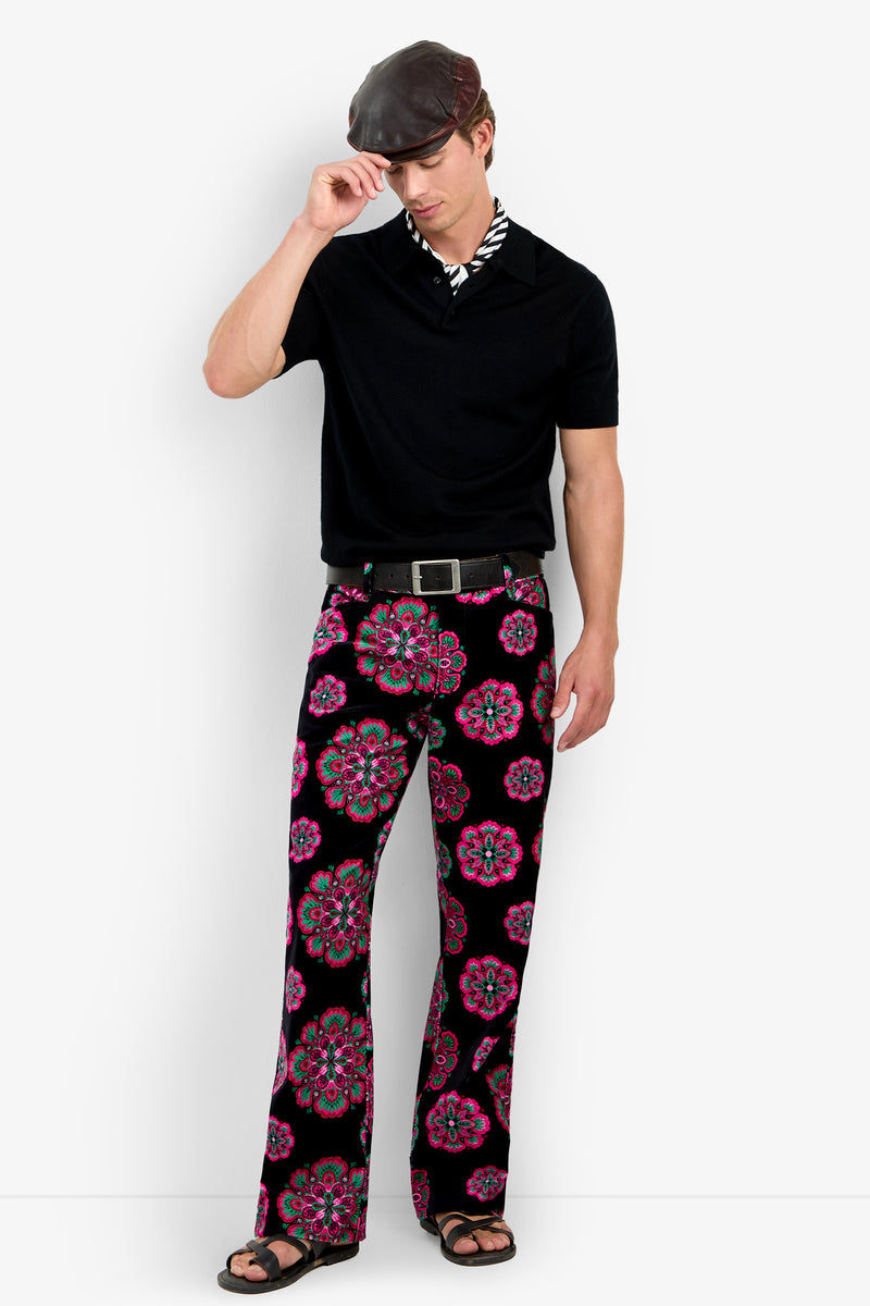 A man wearing a black shirt, the Alejandro Bootleg Trouser with colorful floral patterns, sandals, and a flat cap stands against a white background, touching the brim of his hat with one hand.