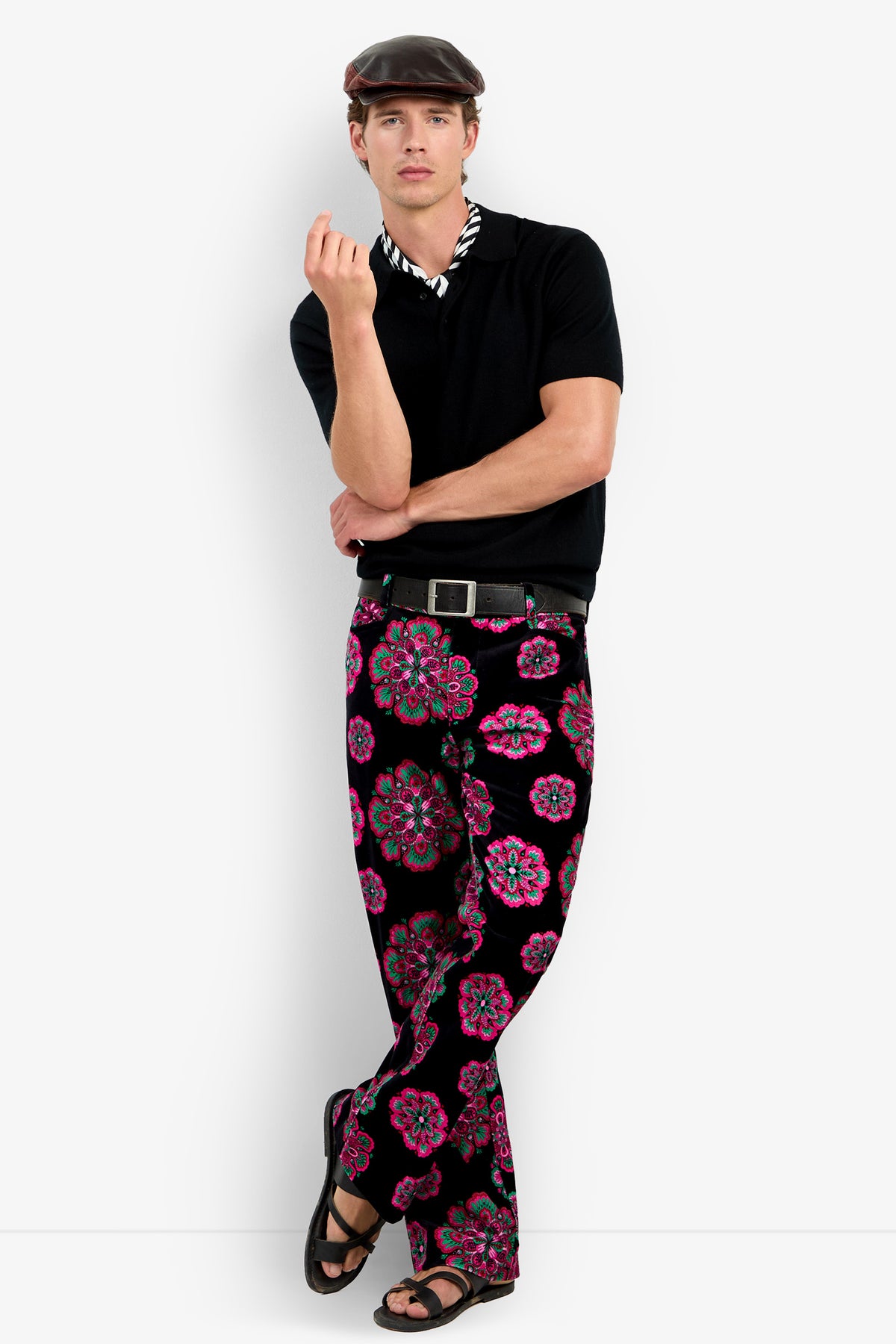 A man stands with one leg crossed, holding his hand up, wearing vibrant Alejandro Bootleg Trousers along with a black shirt, patterned scarf, and maroon cap against a plain white background.